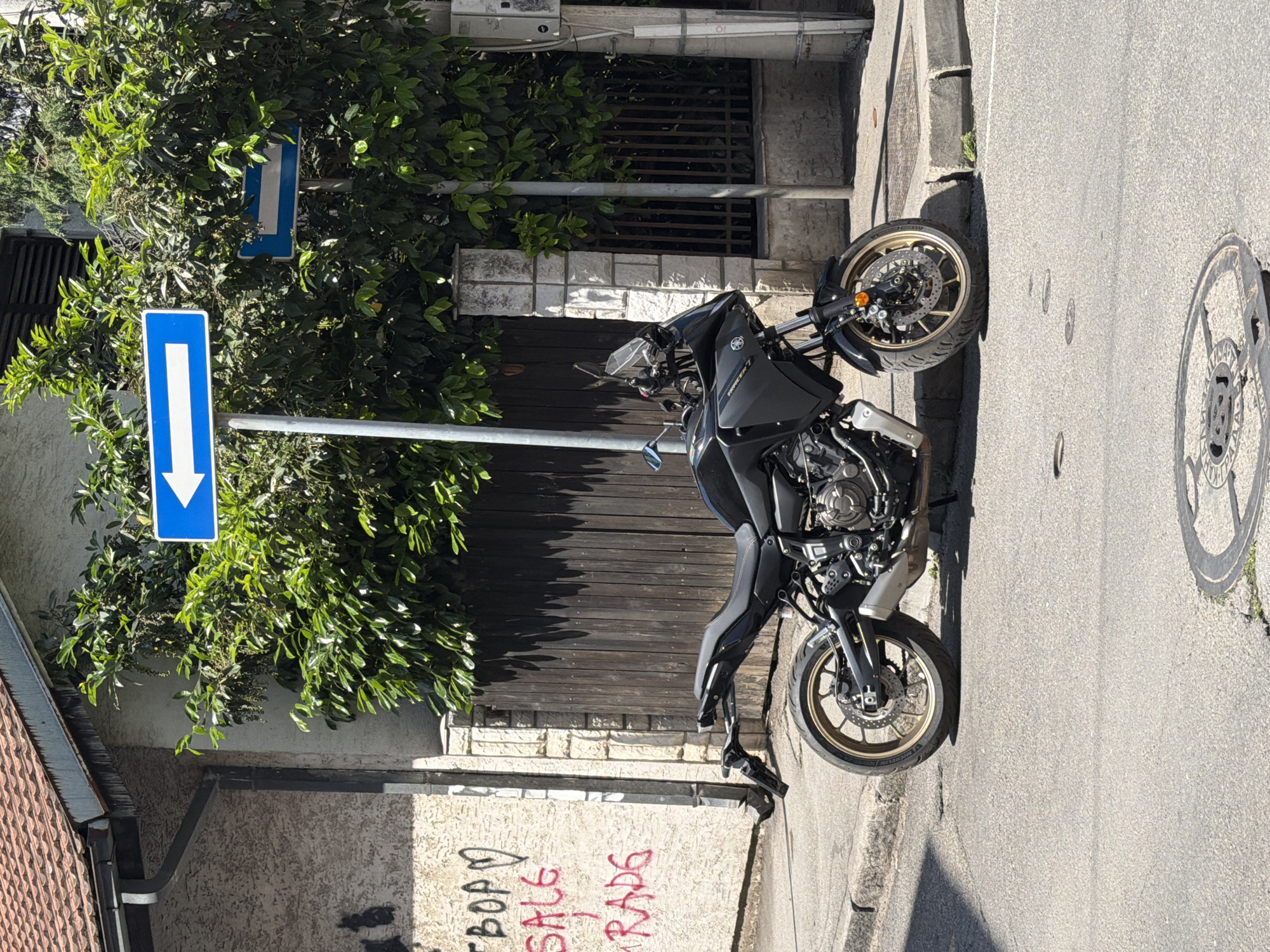 A black motorcycle is parked on the edge of a street in Belgrade, Serbia. The motorcycle is positioned perpendicular to the curb. A blue road sign with a white arrow pointing downwards is visible to the left of the motorcycle, partially obscured by greenery. Graffiti is present on a wall behind the motorcycle that reads "SALE TRADE". A manhole cover is embedded in the asphalt on the right side of the image. The surrounding environment consists of buildings with textured facades and a metal gate. The scene is illuminated by daylight.