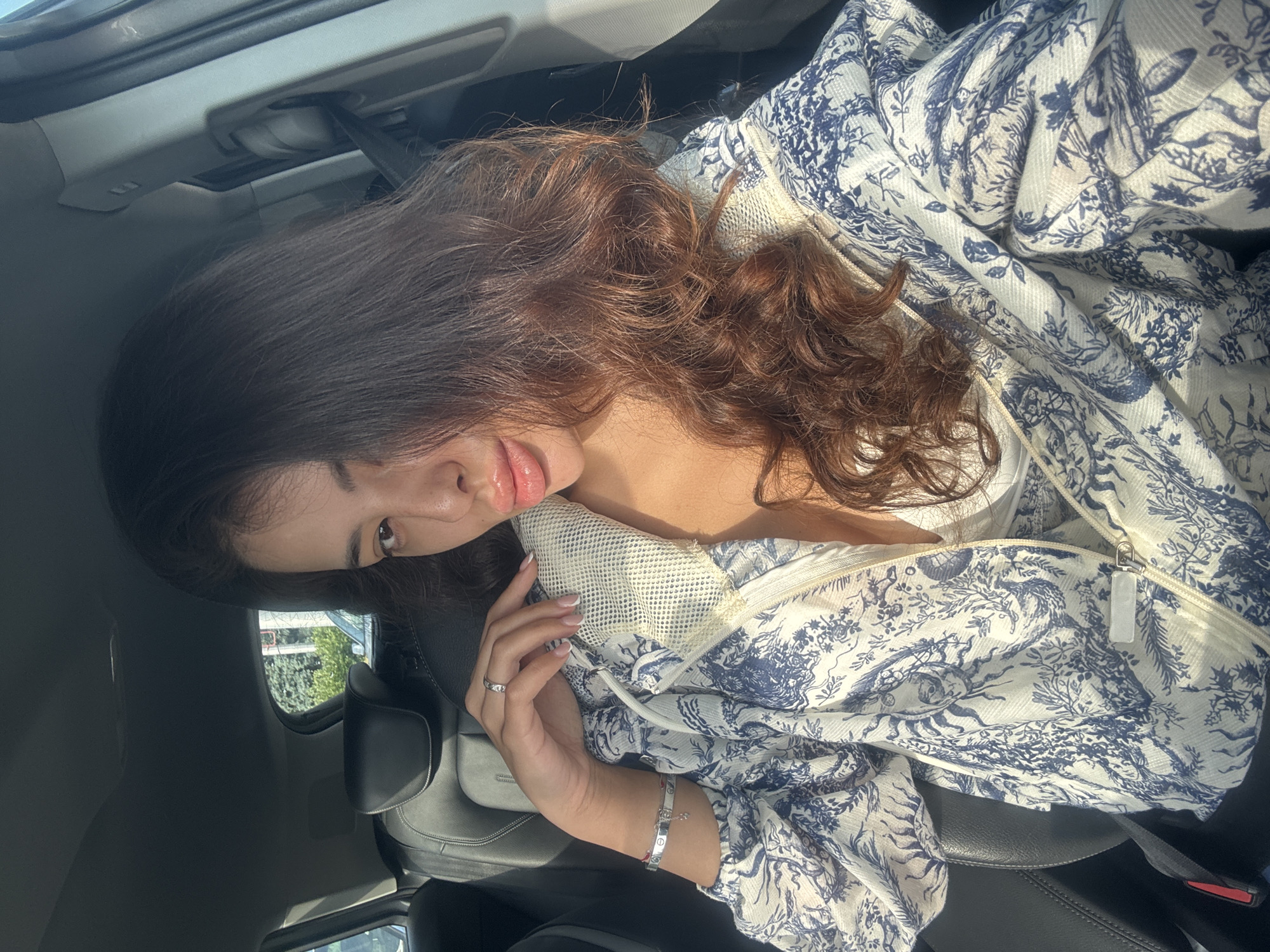 The image shows a woman sitting in the driver's seat of a car. She is looking at the camera, with her right hand resting on her left shoulder. She is wearing a white and blue patterned jacket over a white top. Her hair is long and brown. The interior of the car is visible, including the dashboard and seats. Sunlight is coming in through the windows, creating shadows on her face. The background is blurred, but appears to be an outdoor setting with trees and buildings, suggesting she is parked on a street in the city of Plovdiv, Bulgaria.