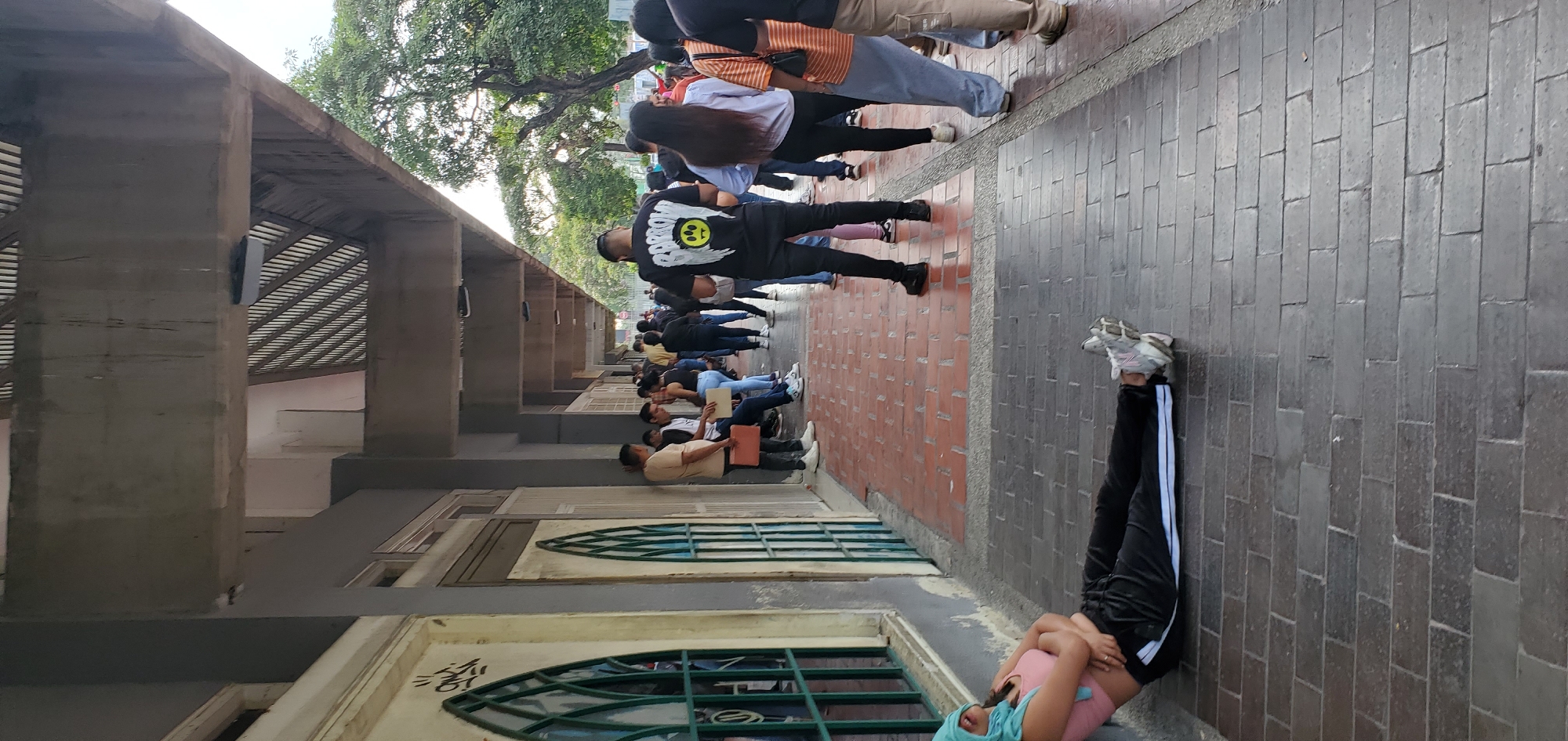 A street scene in Barquisimeto, Venezuela, shows a queue of people lined up against a building. The building has a series of arched windows with ornate metal grilles and a concrete overhang supported by columns. The people are standing or sitting along the brick-paved sidewalk, waiting in line. In the foreground, a person is lying on their back with their legs up against the wall. The street is shaded by trees and the building's overhang.