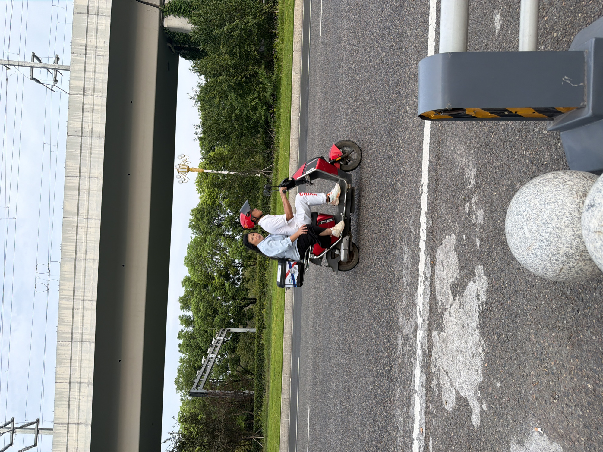 Two people are riding on a red electric scooter on a paved road, likely in Jiangjin, China. The driver, a man wearing a white shirt, white pants, and a red helmet, is operating the scooter. The passenger, a woman in a light blue shirt and black pants, is seated behind him. She is looking towards the camera. A black storage box with a white and blue decal is attached to the rear of the scooter. The scooter has a small front wheel and a larger rear wheel. In the background, a gray overpass with overhead power lines stretches across the frame. Trees and lush green foliage line the side of the road. The sky is overcast. The road surface has white lane markings and some lighter colored patches of unknown origin. In the foreground, a portion of a gray barrier with yellow and black hazard stripes is visible, along with two round, speckled gray objects.