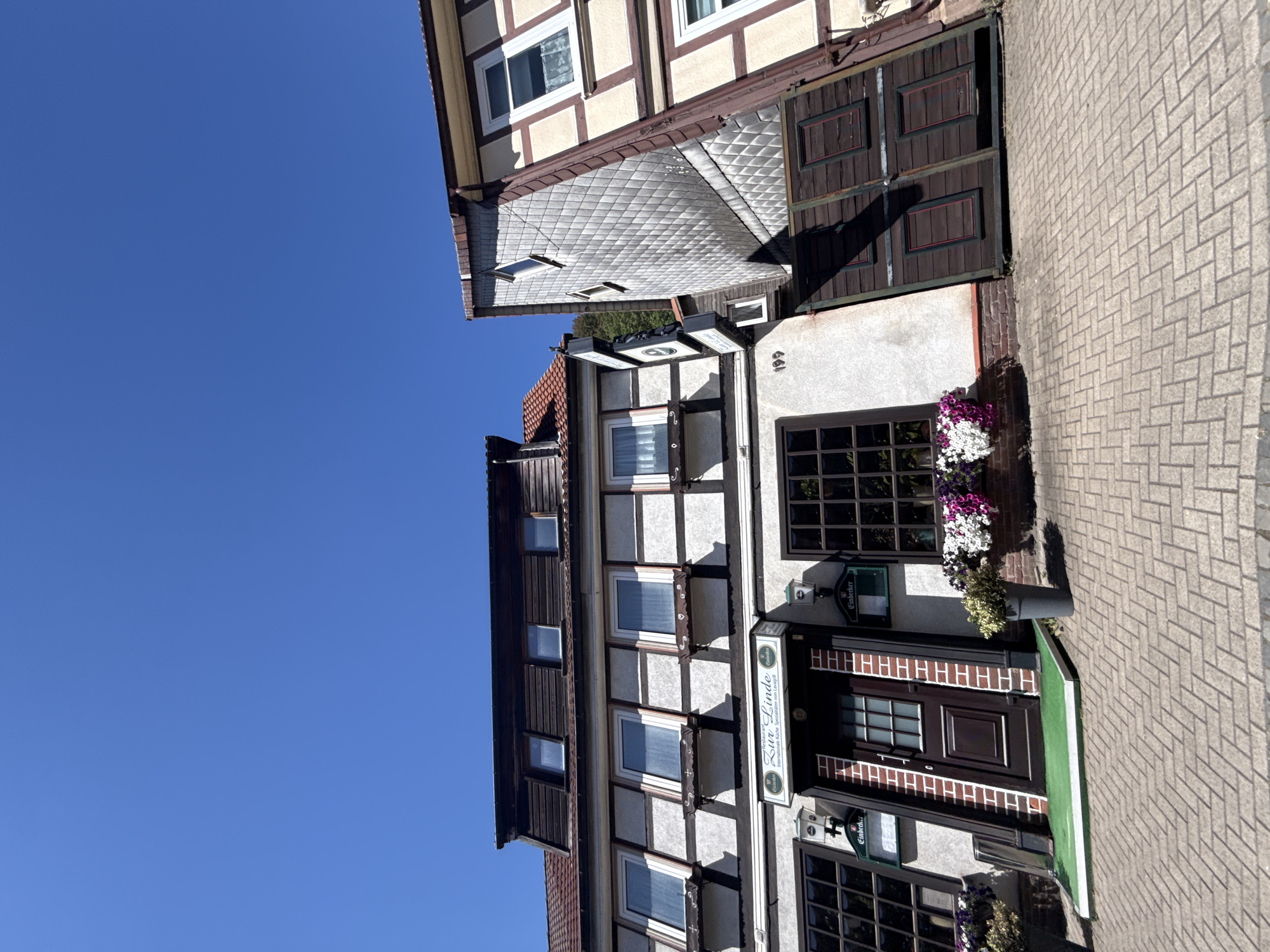 A wide-angle, low-angle exterior shot captures a multi-story half-timbered building under a clear blue sky. The building appears to be a restaurant or inn, as indicated by signage that reads "Restaurant Zur Linde". The facade of the building features exposed dark timber beams against a lighter plaster. Several windows with white frames and dark pane detailing are visible on multiple levels. Some windows have flower boxes with colorful blooms. A dark brown door with a glass-paned upper section is centrally located on the ground floor, accessible by a small green carpeted ramp. Potted plants are placed on either side of the entrance. The foreground is paved with light-colored bricks laid in a herringbone pattern. To the right of the main building, another section of the structure, also half-timbered, is visible, with a distinctive tiled facade. The overall scene suggests a well-maintained establishment in a town, likely in a historic district, given the architectural style. The location is identified as Bad Lauterberg im Harz, Germany.