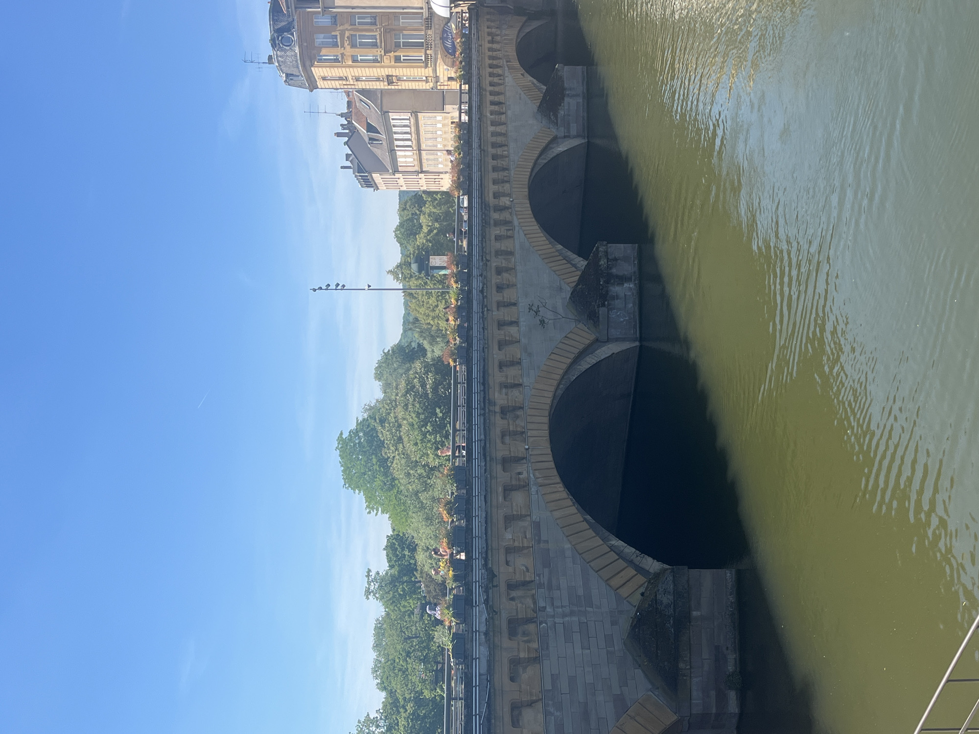 This outdoor shot, taken in Metz, France, displays a portion of a stone bridge traversing a river. The bridge features multiple arches visible above the water line. Adjacent to the bridge are buildings of varying heights, a line of trees, and a lamppost with perched birds. The river is greenish-yellow, reflecting the surrounding environment. The sky is clear and blue.