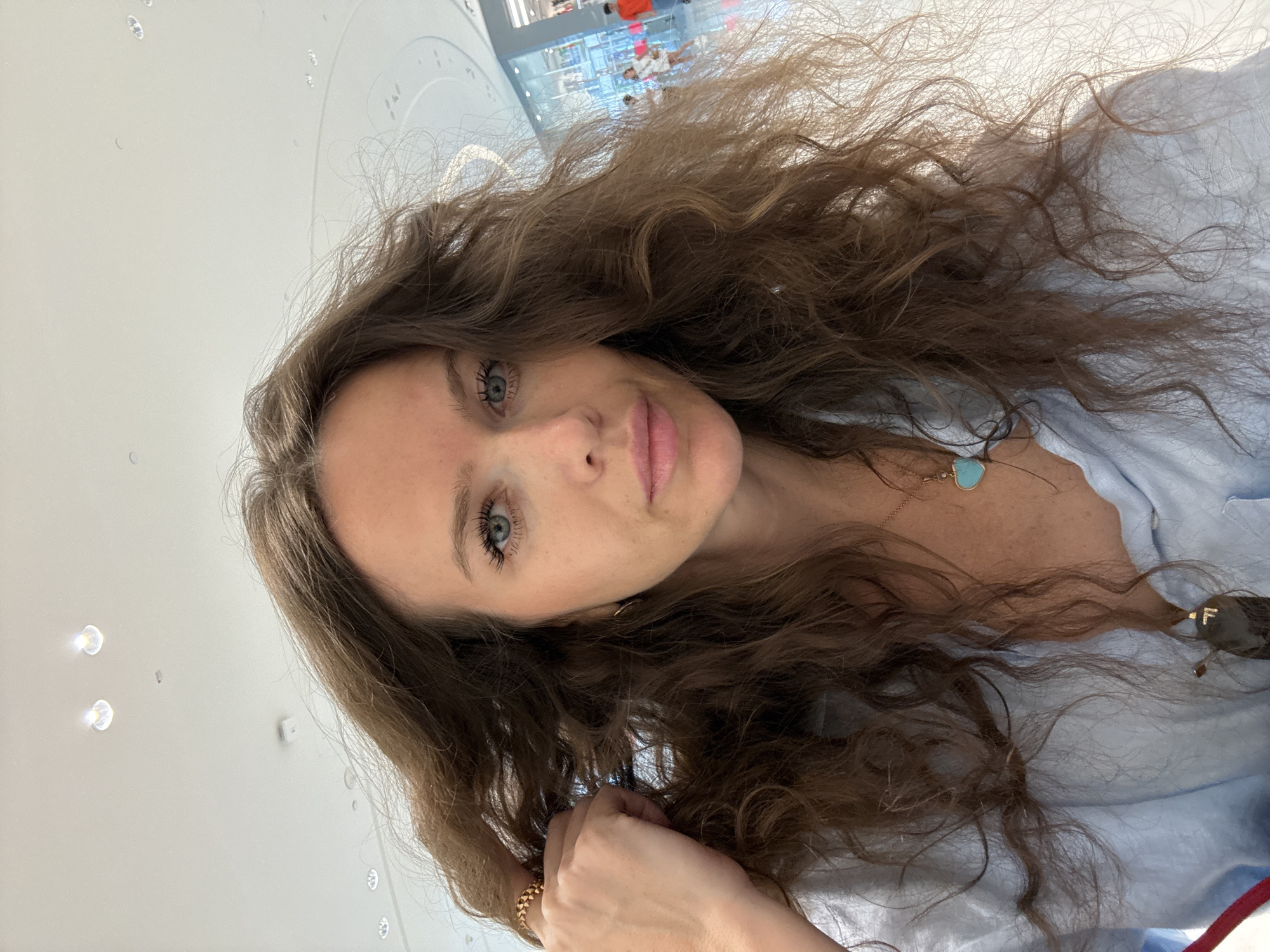 A medium shot selfie of a woman with long, wavy brown hair. She has blue eyes and is wearing a light blue shirt and a gold necklace with a turquoise heart pendant. Her left hand is raised and touching her hair. She is wearing a ring on her left index finger. The background appears to be a brightly lit indoor space, possibly a shopping mall, with a reflective floor and a high ceiling with recessed lighting. In the distance, there are shops and other people. The overall lighting is bright and natural.