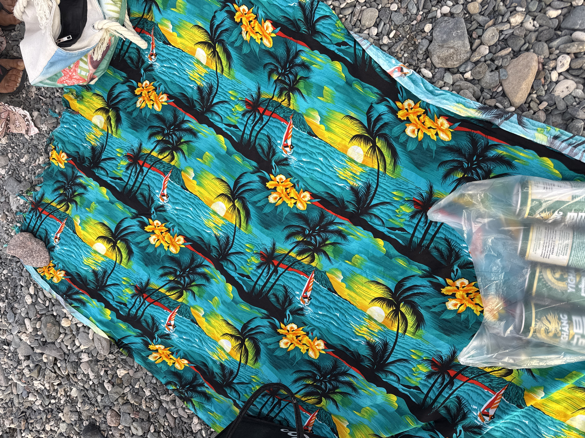 A brightly colored Hawaiian-themed beach towel is spread on a rocky shore. The towel features a pattern of palm trees, yellow flowers, and red and white surfboards on a turquoise and yellow striped background that evokes water and sunlight. In the lower portion of the image, a clear plastic bag contains several cans of beverages, some with visible labels including "CHANG" and "YICHANG." A woven bag with a zipper and rope handle rests on the right side of the towel. The ground is covered in various sizes of gray and brown pebbles and stones, characteristic of a beach or riverbed. The scene appears to be outdoors. There are no visible people actively participating in any activity. The location is identified as Urdzhar, Kazakhstan.