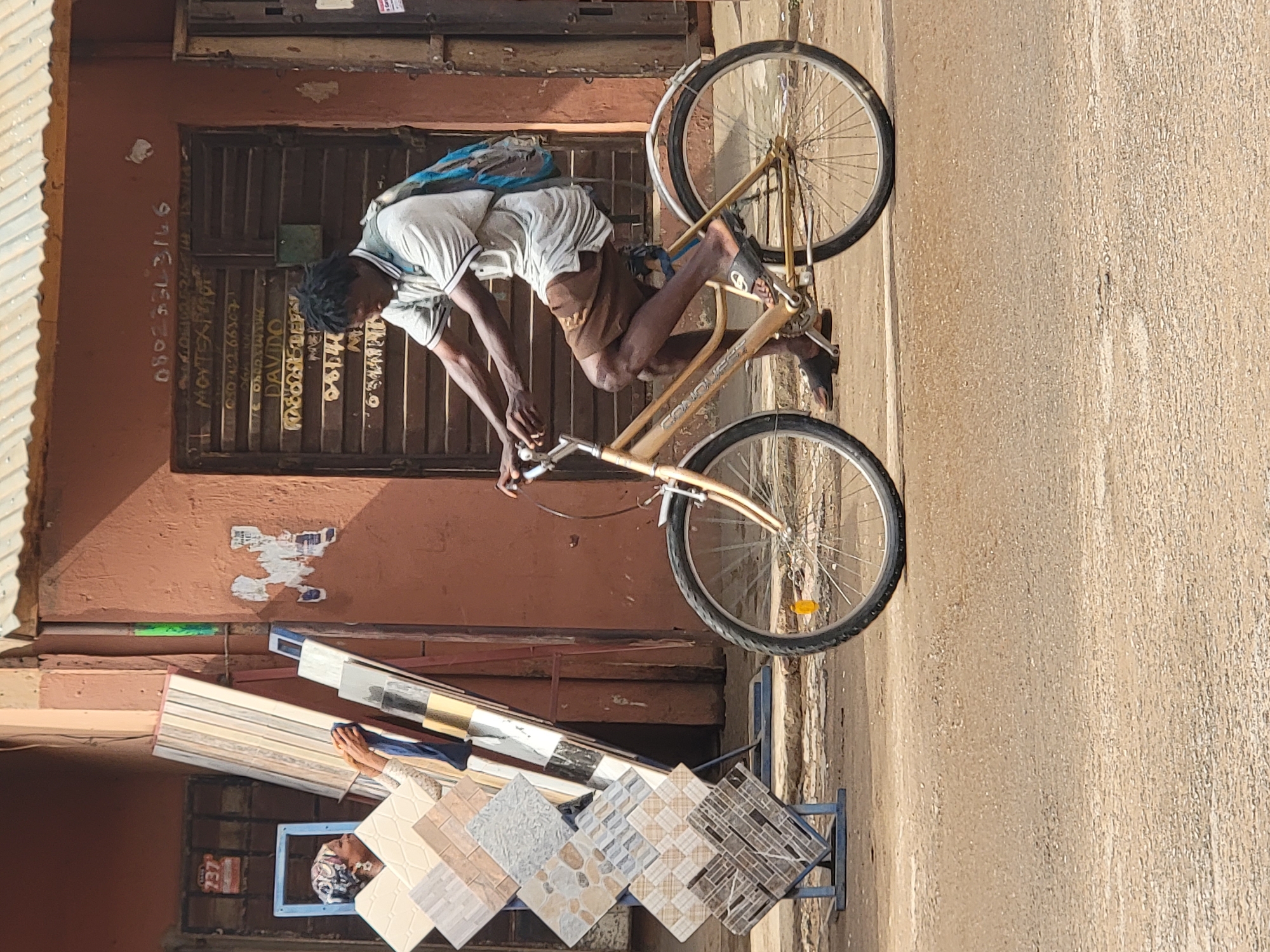 In Maiburiji, Nigeria, a man rides a gold bicycle on a street. He is wearing a white shirt and brown shorts, and has a blue bag on his back. He is passing in front of a building with a closed brown shutter door. A display of square tiles sits on a blue frame in front of the building. The street is paved.