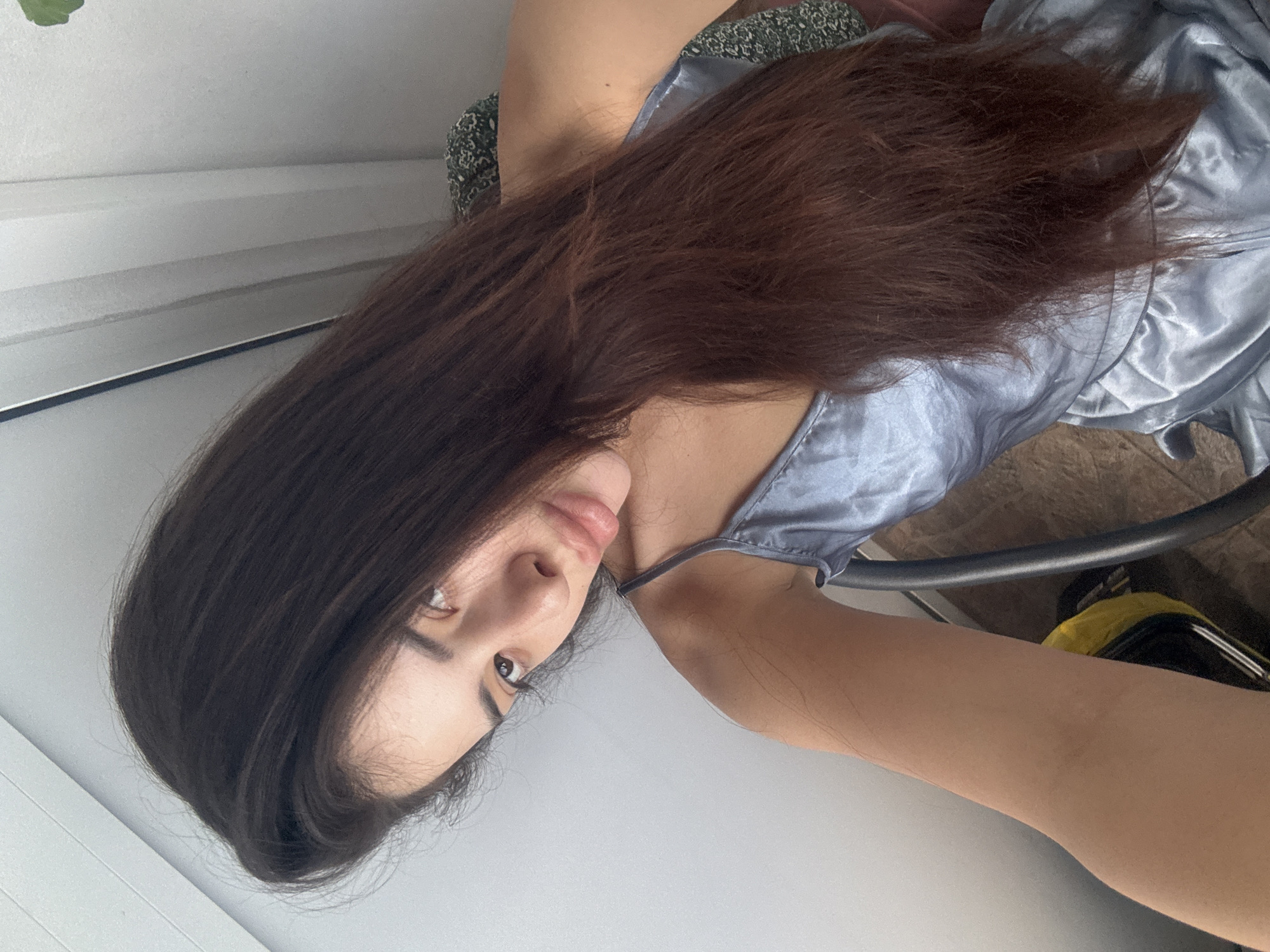The image, oriented counter-clockwise, depicts a young woman in an indoor setting. She is likely reclining or lying down, with her long, dark brown hair fanning out around her. She wears a light blue or silver satin slip dress with thin straps. Her head is tilted, and she gazes with a soft, somewhat pensive expression, her dark eyes looking towards the camera. Her facial features include full lips and a delicate nose.
Partially visible behind her shoulders is a dark green patterned fabric, possibly from a cushion or chair. A black metal chair component and a hint of a yellow object are seen towards the bottom right. The background is a plain, light-colored wall, possibly featuring horizontal elements resembling window blinds or wall panels on the left. The scene is brightly and evenly lit, suggesting it is daytime. No text is present in the image. The location context for this media file is Plovdiv, Bulgaria.