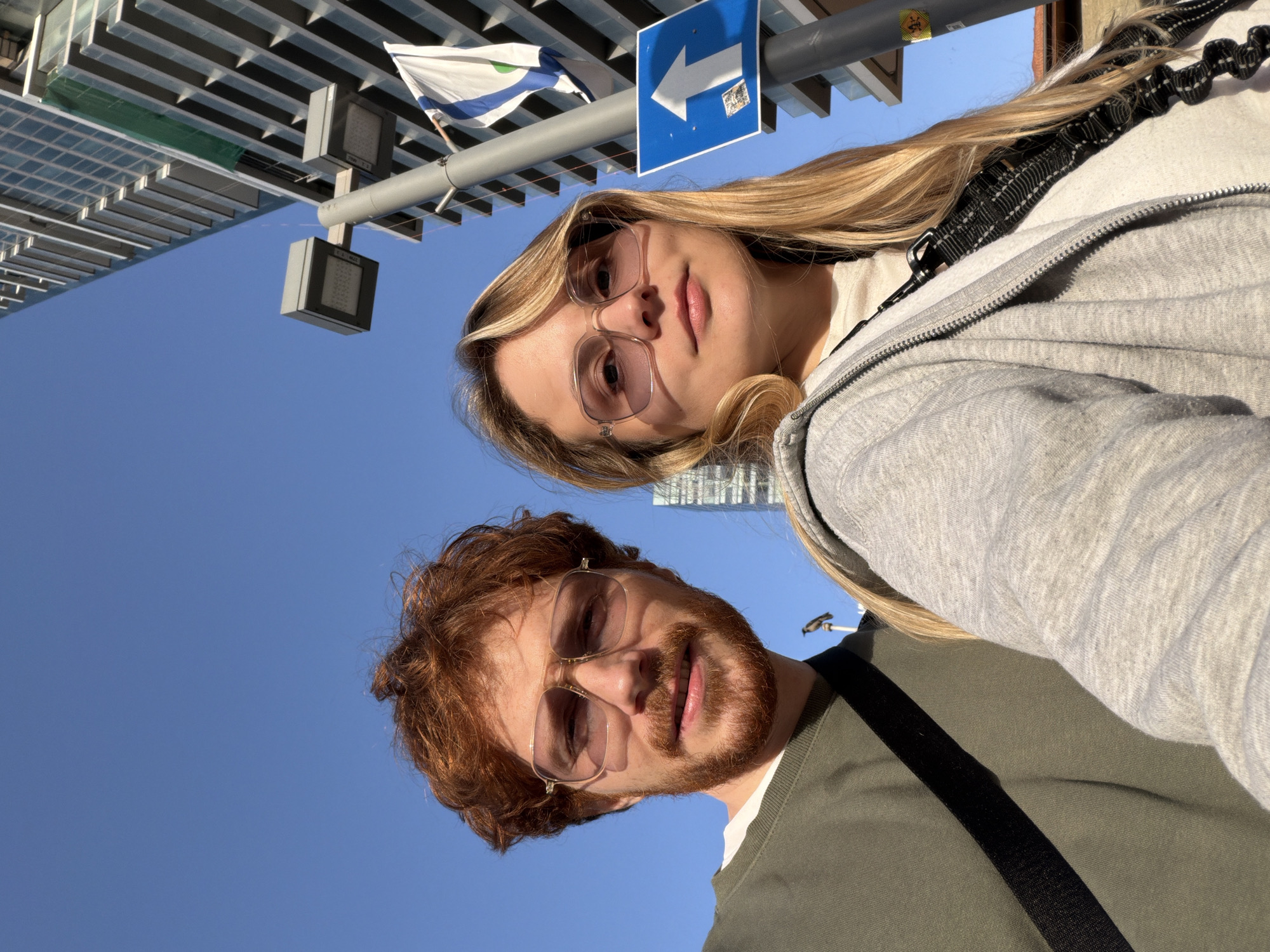 Two individuals, a female and a male, are oriented in a selfie taken at a tilted upward angle against a clear blue sky. The female has long blonde hair and wears transparent-framed glasses and a grey zip-up hoodie. A black strap is visible across her torso. The male has red hair and a beard, wears transparent-framed glasses, and is dressed in an olive green shirt.
Background elements include a directional blue and white street sign pointing left, a white flag featuring blue stripes and a green triangle, and a modern building with a grid-like facade. The location is in Tel-Aviv, Israel.
