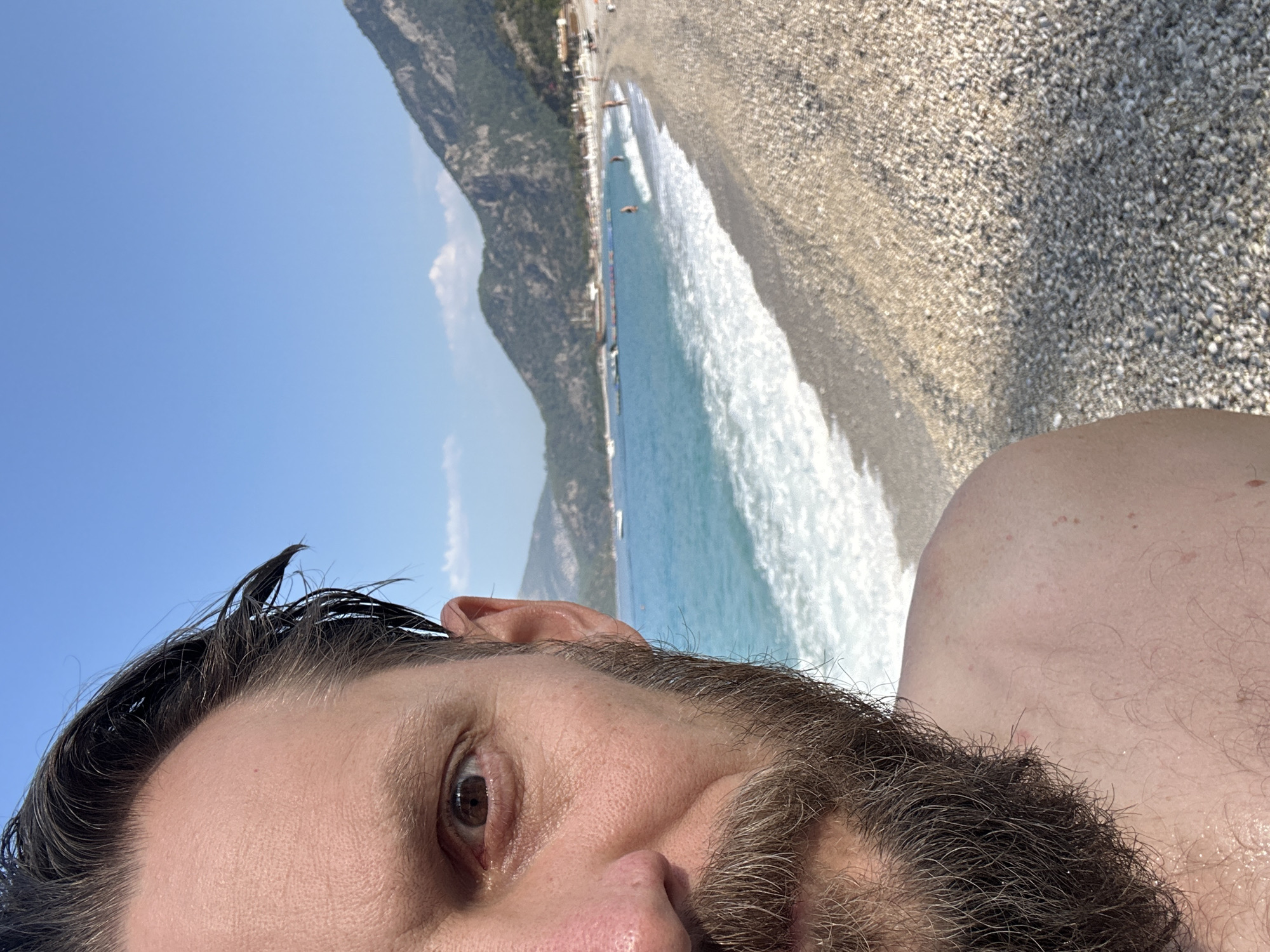 The image is a close-up, selfie-style shot of a man's face and shoulder, taken on a beach in Fethiye, Turkey. The man has dark hair, a beard, and is looking towards the camera with an open eye. His skin appears slightly damp, suggesting he may have been swimming. The background shows the turquoise waters of the Mediterranean Sea, a pebbled beach, and a coastline with mountains. The sky is clear and blue with a few scattered clouds. Several people are visible in the water and on the beach in the distance, engaged in leisure activities. A boat is also present in the sea. The overall setting is a sunny day at a popular tourist destination.