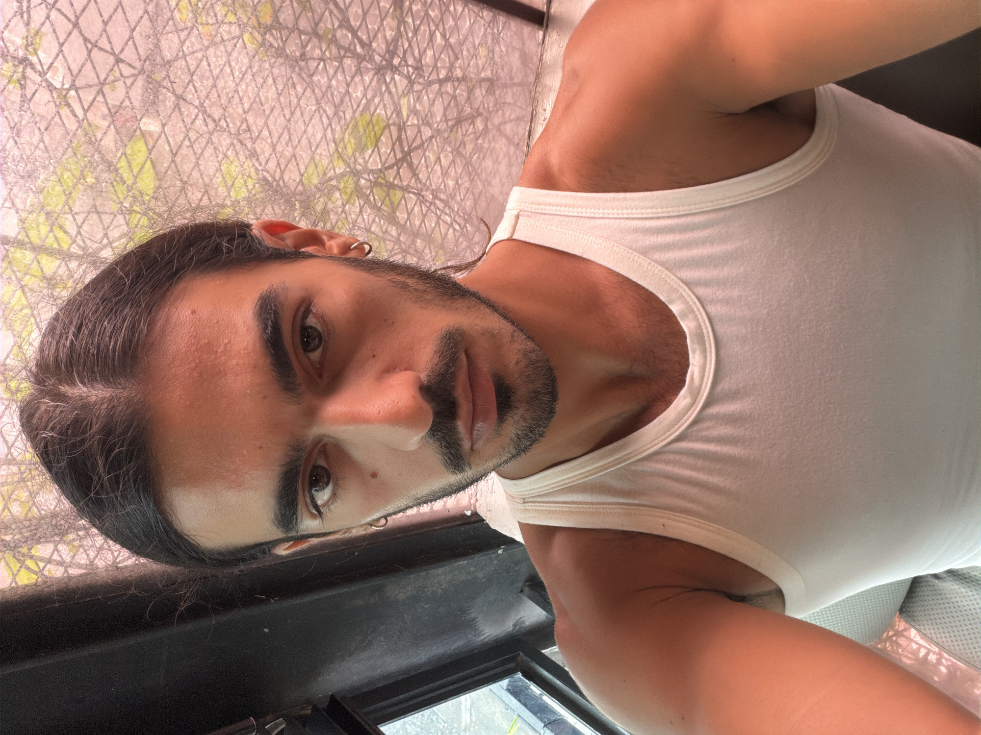 A vertical selfie shows a young adult man looking directly into the camera with a neutral expression. He has dark, long hair parted down the middle and pulled back, a short beard, and a mustache. He wears a plain white tank top and a silver hoop earring in his visible left ear. The background features a light-colored, diamond-patterned mesh screen, possibly a fence or decorative panel, through which blurry green foliage is visible. Below his face, a dark, rectangular structure, possibly a window frame or railing, is present. A light grey mesh texture, potentially a seat or cushion, is partially visible under his right arm. The lighting suggests it is daytime. The image was taken in North Kuta, Indonesia. No text is visible.