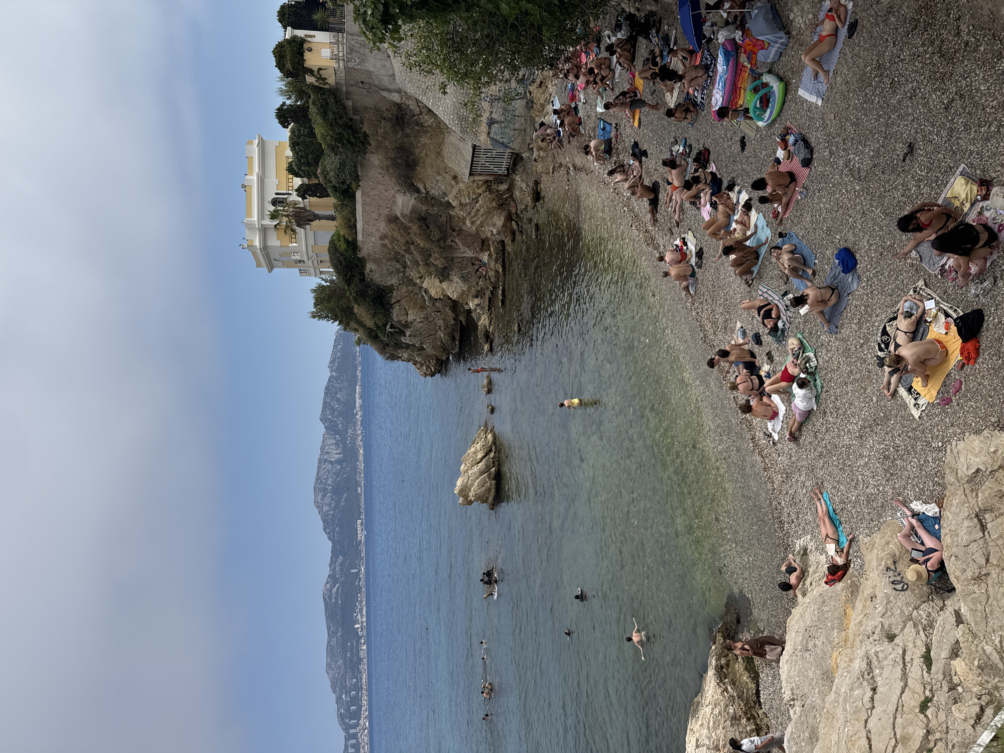 This media file shows a crowded pebbled beach in Marseille, France, with many people sunbathing, swimming, and relaxing. The scene is set on a sunny day with clear blue skies, suggesting it's likely midday or early afternoon during the warm summer months.
The beach is nestled against a rocky cliff face, with a building visible on the upper edge of the cliff. The water is clear and calm, with several people swimming and wading in the shallows. Some individuals are lying on towels and blankets, enjoying the sun, while others are socializing in groups. There are also inflatable toys and beach accessories scattered across the beach.
In the background, a mountainous coastline stretches along the horizon, characteristic of the Marseille region. The overall atmosphere appears relaxed and joyful, with people embracing the outdoor leisure activities. No visible text is present in the media file.