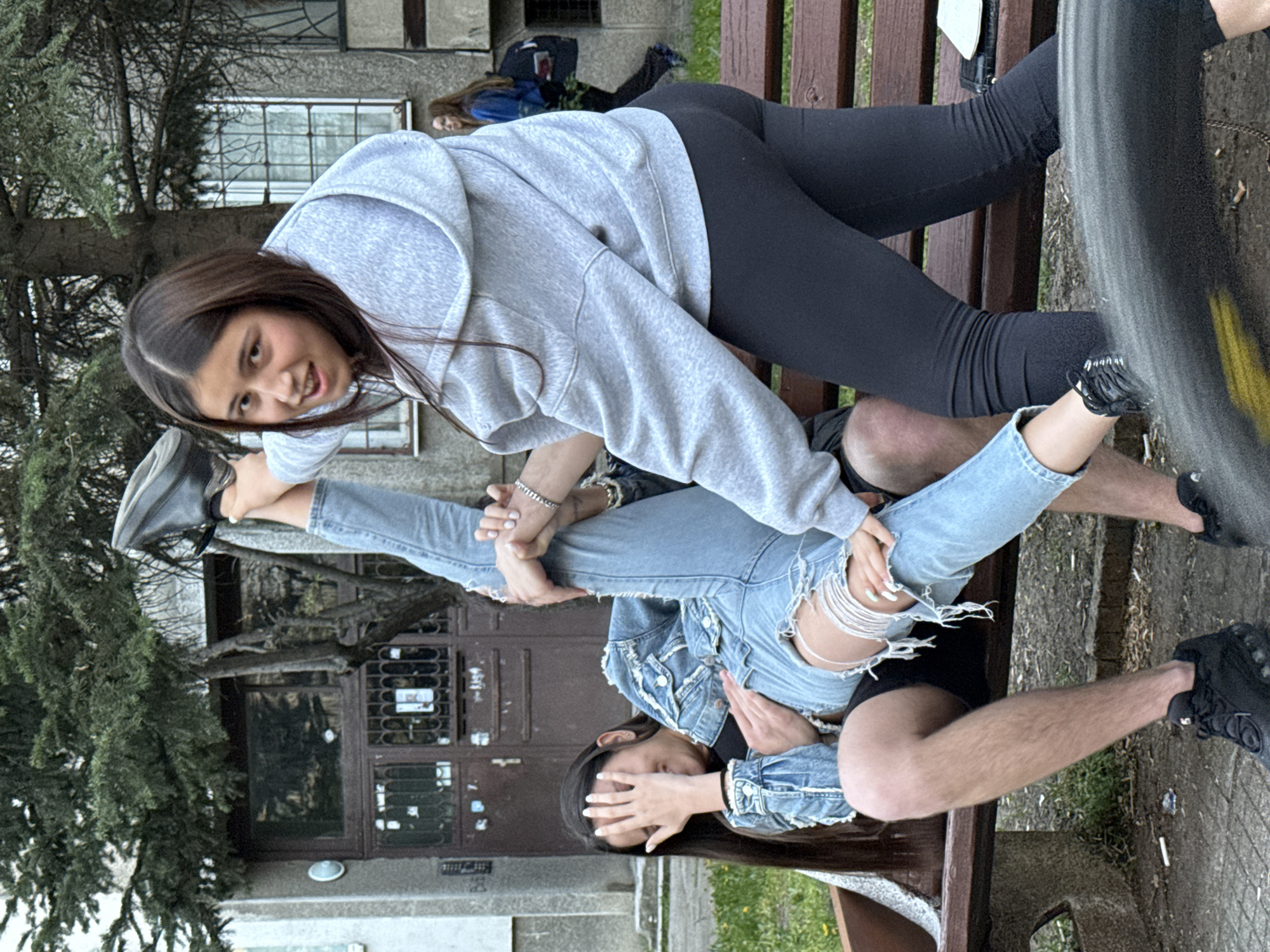 Two young women are on a bench in front of a building. One woman is lying on the bench with her legs spread apart. She is wearing ripped jeans and a denim jacket. The second woman is standing over her, stretching her leg up. She is wearing leggings and a gray hoodie. The scene appears to be in Sofia, Bulgaria, due to the architectural style of the building in the background. The building is a multi-story brick structure with barred windows and a locked door. There are also several trees around the building and bench.