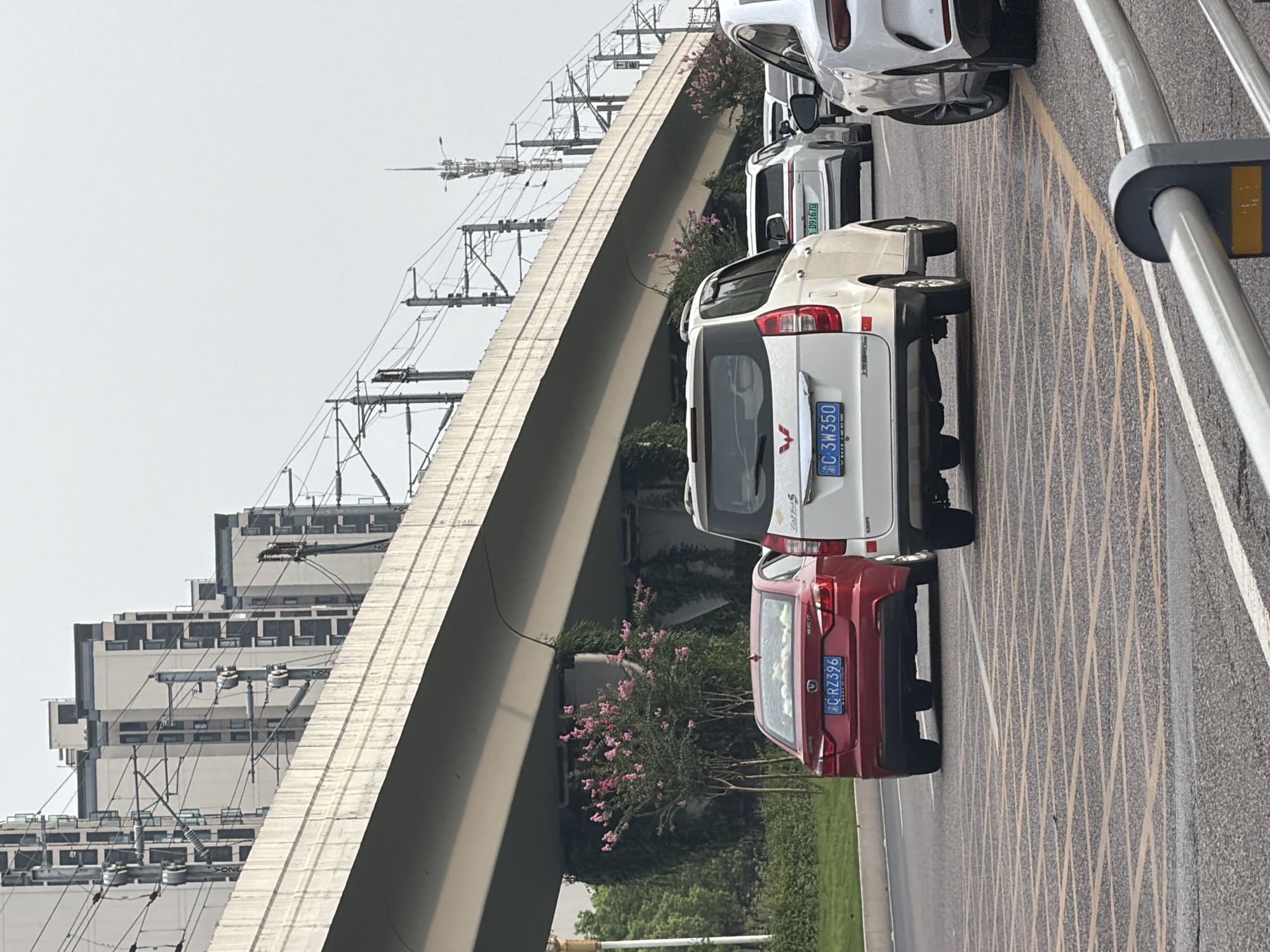 In Jiangjin, China, a traffic scene unfolds beneath an elevated structure that appears to be part of an overpass or railway. The overpass is supported by concrete pillars, and on top of it, numerous electrical wires and pylons are visible, suggesting a transportation corridor. Buildings with a modern, multi-story design are situated in the background behind the overpass. The road below is paved with asphalt and marked with yellow and white lines, indicating lanes and traffic divisions. Several vehicles are present on the road, including a red sedan with license plate "C RZ396", a white minivan with license plate "C 3W350", and another white car. Pink flowering trees are growing beneath the overpass, adding a touch of natural color to the urban environment. The sky is overcast, casting a diffused light on the scene.