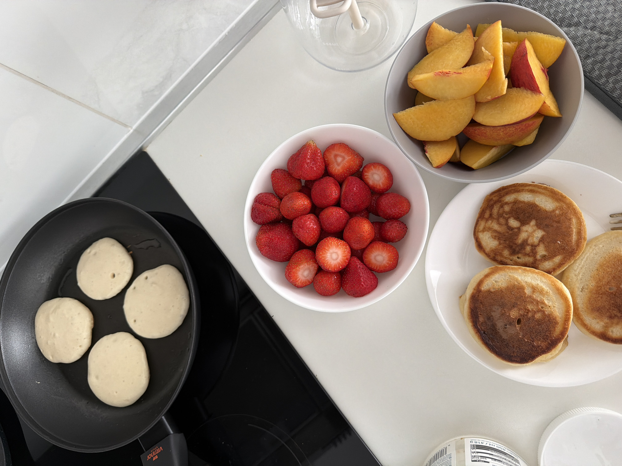 The image depicts a kitchen counter with breakfast items. In a black frying pan on a black induction stovetop, four pancakes are cooking. Next to the stovetop, on the counter, are three white bowls. The first bowl contains sliced strawberries. The second bowl contains sliced peaches. A white plate holds three cooked pancakes. To the left of the plate is a white jar with a white lid. The background shows a white countertop with a marble-like pattern. There is no identifiable location in Finestrat, Spain.