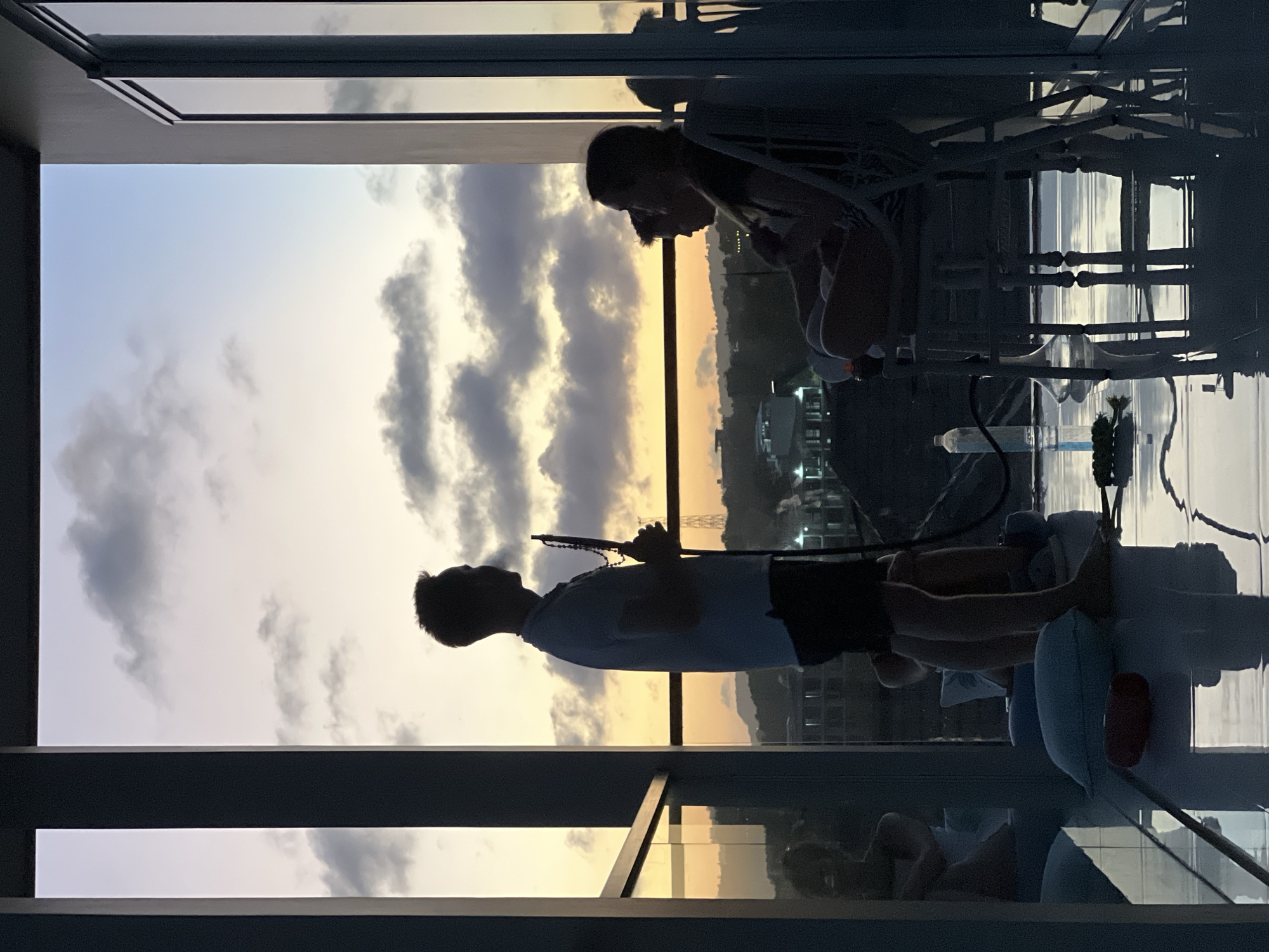 Two individuals are present on a balcony, with the city skyline visible in the background. One person is standing and appears to be holding rosary beads, while the other is seated. A hookah is also visible. The location appears to be a high-rise building or apartment in an urban area. The lighting suggests it's either sunrise or sunset.
