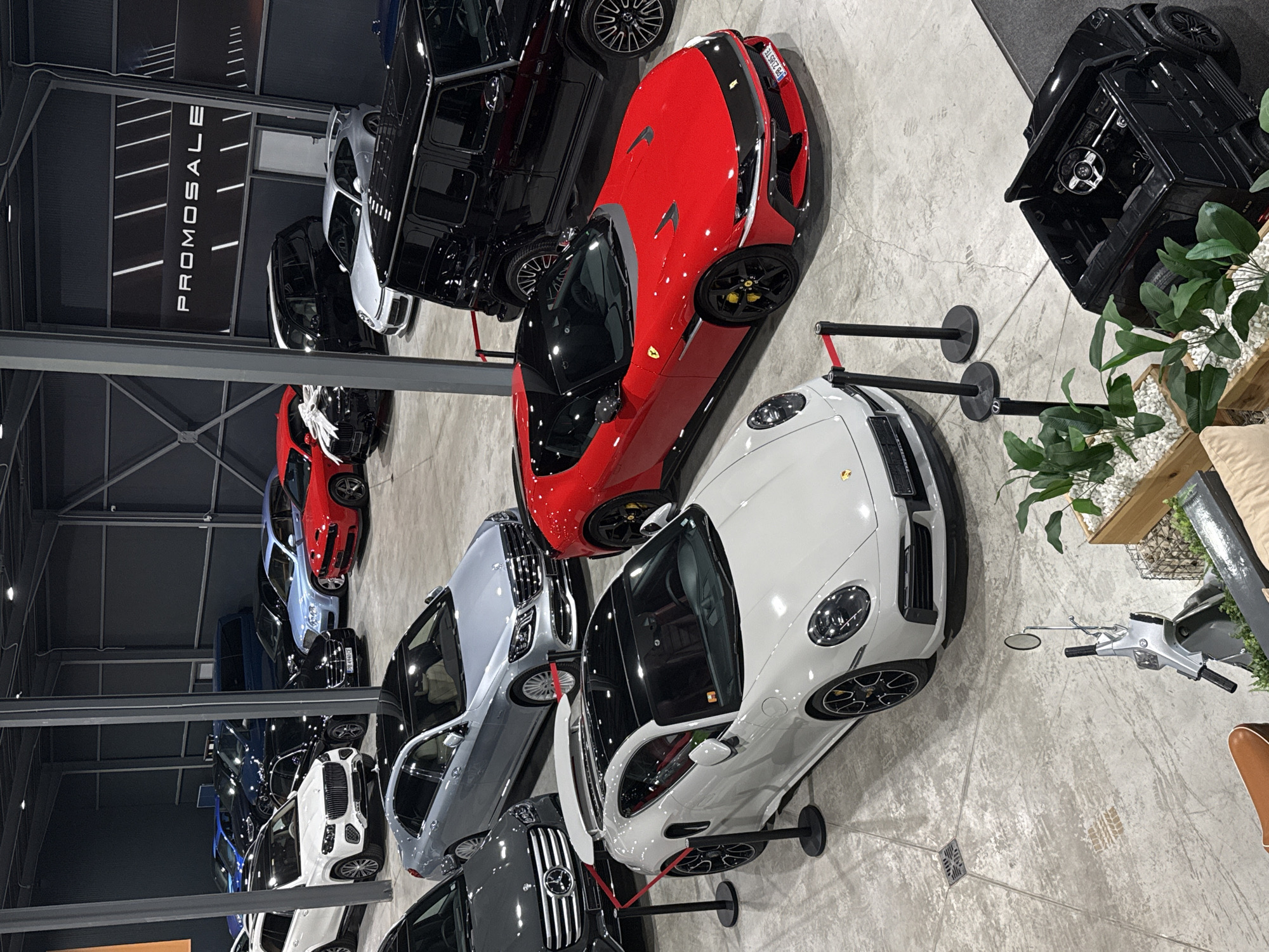 This image captures an indoor car showroom in Plovdiv, Bulgaria, from an elevated perspective. The scene showcases a variety of luxury and performance vehicles displayed on a concrete floor with visible tire marks and scuffs.
In the foreground and center, a vibrant red sports car, possibly a Ferrari or similar model, is prominent, featuring black wheels with yellow brake calipers and a license plate reading "PB 2045 TC". Adjacent to it, a sleek white Porsche 911-like sports car with black multi-spoke wheels is partially visible, demarcated by black stanchions with red retractable belts. Behind the red car and to the left, a large black SUV, resembling a Mercedes-Benz G-Wagen, is parked. Further left, a silver sedan, likely a Mercedes-Benz, can be seen.
The showroom features dark grey walls and metal structural beams. A large illuminated sign on a dark wall clearly displays the word "PROMOSALE", indicating a sales event. In the background, numerous other cars, including black, white, and blue models, both SUVs and sedans, are neatly arranged, some with decorative white bows on their hoods.
To the right of the white sports car, a small black ride-on toy car resembling an SUV is parked next to a wooden planter filled with green plants and white decorative stones. A silver scooter or moped, similar to a Vespa, is also visible near the planter. Part of a brown piece of furniture can be seen in the bottom right corner. The overall lighting is artificial, illuminating the cars and the display area. No people are present in the image.