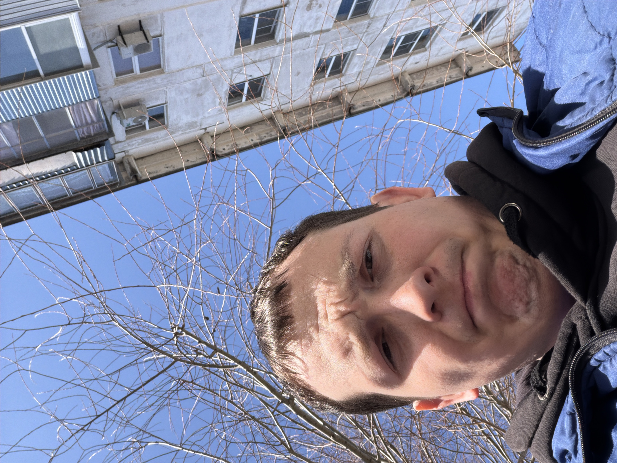 A man with a furrowed brow is photographed from a low angle against a backdrop of bare tree branches and a multistory building under a clear blue sky. The building appears to be of Soviet-era construction, with multiple windows and air conditioning units mounted on the exterior. The man is wearing a dark hooded sweatshirt and a blue jacket. The location is Chaikovsky, Russia.
