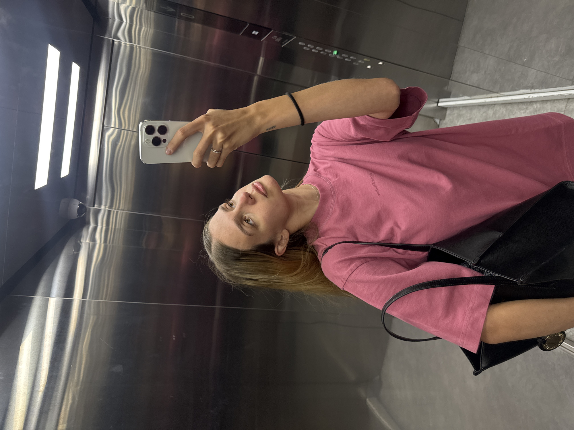 A person is taking a selfie in an elevator. The elevator has stainless steel walls and bright, vertical lights. The person is wearing a pink t-shirt and holding a white smartphone. A black bag hangs on their shoulder.