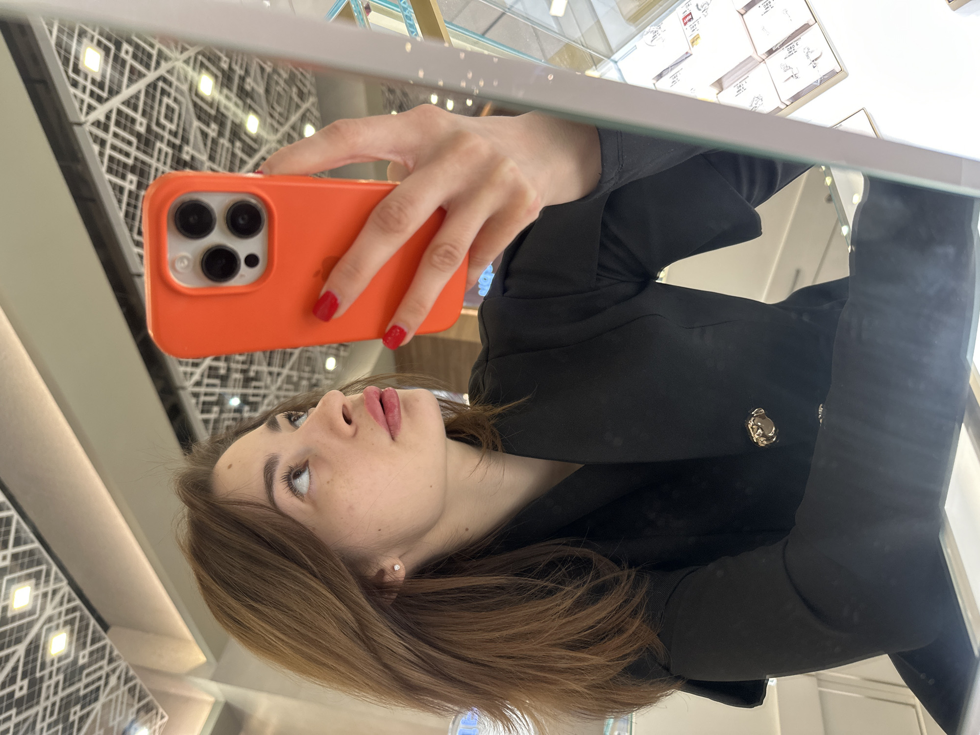 The image is a mirror selfie taken by a woman in the Greenwich Shopping Center (ТЦ Гринвич) in Yekaterinburg, Russia. The woman, with medium-length brown hair, light skin, and freckles, is depicted from the mid-torso upwards, looking slightly upwards and to her right. She wears subtle makeup, including a pinkish lip color and mascara, and small stud earrings. Her attire is a black blazer or jacket featuring a single visible golden, ornate button. She holds an orange-cased smartphone, identified as an iPhone with three prominent rear camera lenses, in her right hand, which displays red-painted fingernails. The background, visible in the mirror reflection, features a distinctive geometric ceiling pattern composed of dark lines forming a grid of diamonds and squares against a lighter surface, with integrated rectangular light fixtures. To the woman's right, a portion of a glass display case with various small white boxes or items and what appear to be price tags is visible, indicating a retail setting likely for jewelry or small luxury goods. The environment is brightly illuminated.