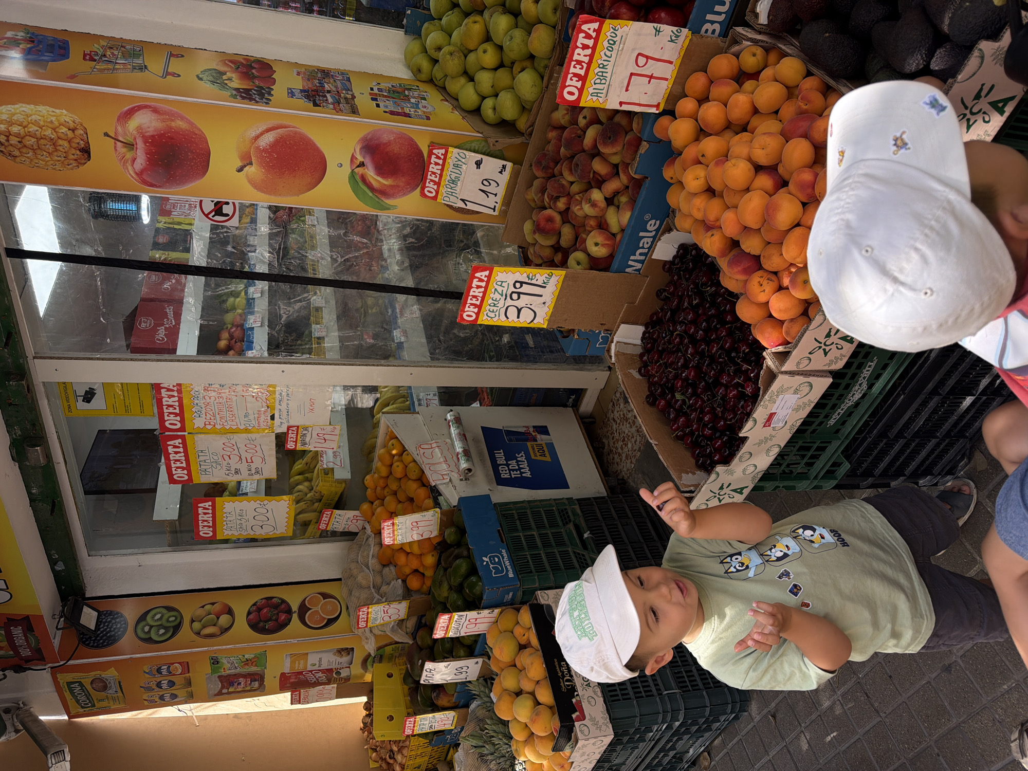 The image shows a fruit stand located outdoors. A young boy wearing a light green t-shirt and a white baseball cap is looking at a piece of dark fruit in his hand. He is standing in front of various crates filled with fresh produce, including cherries, apricots, and other fruits. A second child, wearing a white baseball cap and a red and white striped shirt, is partially visible in the foreground, facing away from the camera. The fruit stand displays various "Oferta" (offer) signs with prices written in Euros. In the background, the interior of a shop is visible, with shelves stocked with packaged goods. The overall environment suggests a market or a street-side vendor.