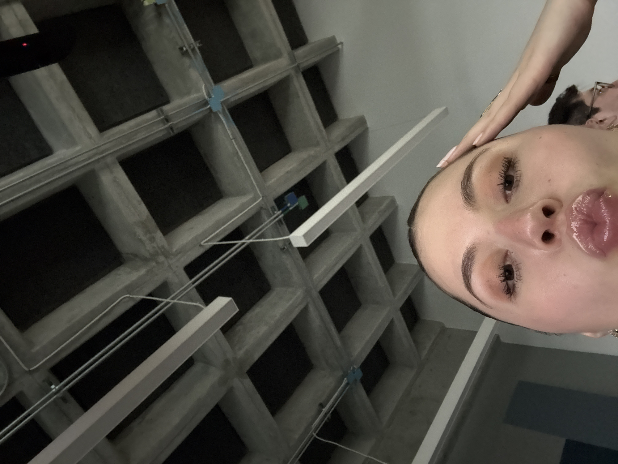 The image shows a person's face, likely taken with a smartphone at a low angle. Another person's hand is on their forehead, and part of another person's head is visible in the background. The background features a concrete waffle slab ceiling with electrical conduit and lighting fixtures. Based on the architectural style, this ceiling is commonly found in buildings in Mexico City, Mexico.