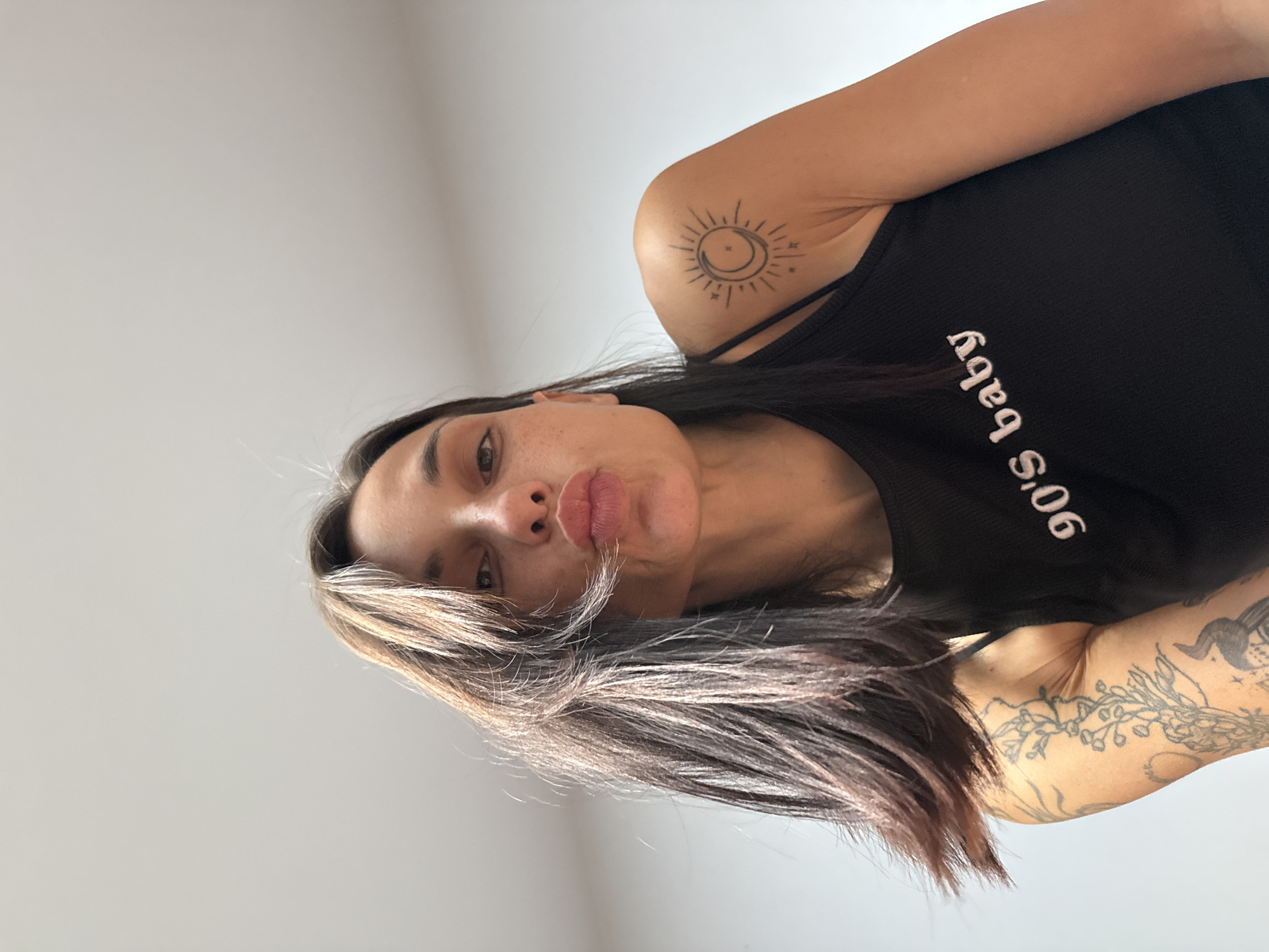 In the city of Pleven, Bulgaria, a woman with dark hair and fair skin is pictured from a low-angle perspective. She is wearing a black tank top with the text "90's baby" printed on it in white. She has tattoos on her right shoulder, depicting a sun and moon, and on her left arm, featuring floral designs and other imagery. The woman is puckering her lips as if for a kiss. The background is a plain, light-colored wall, suggesting an indoor setting. The lighting appears to be natural, possibly from a window.