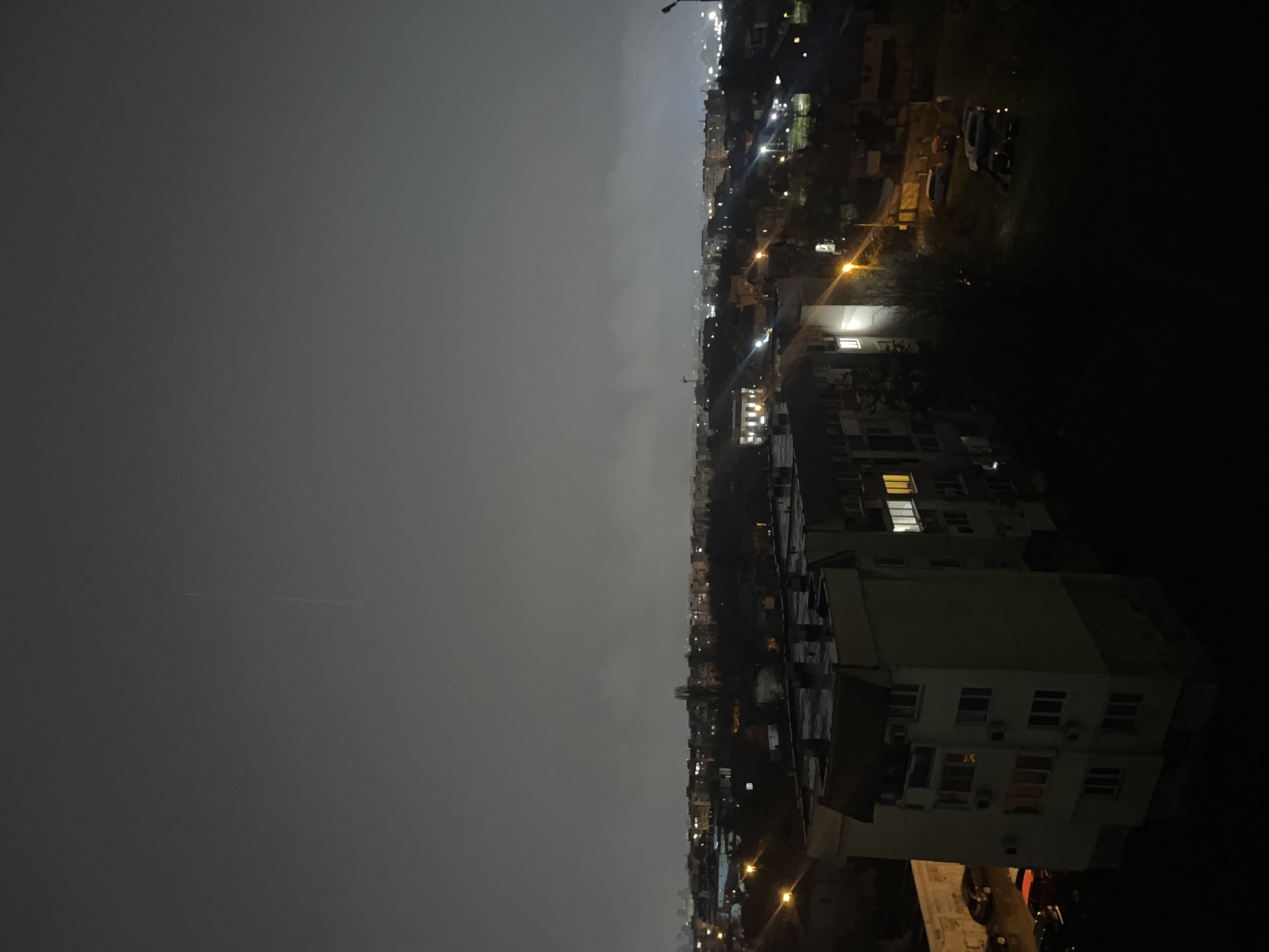 This nocturnal image captures a dense urban residential district in Sofia, Bulgaria, viewed from an elevated vantage point. A thick layer of low-lying cloud or fog obscures the sky and diffuses all artificial light sources, with a faint, thin vertical streak visible in the upper-middle atmosphere.
In the immediate foreground, a light-colored, five-story apartment building with a flat roof and multiple exterior air conditioning units occupies the lower left. Several windows within this building emit warm yellow and white light. Adjacent to its base, a dark street or parking area holds multiple parked vehicles, including a red car and a dark sedan, positioned next to a low concrete wall.
The midground presents a sprawling urban expanse filled with numerous residential blocks. Streetlights and illuminated windows throughout the district cast diffused light. Bare trees are scattered among the buildings. To the right, a prominent streetlamp with a bright yellow emission illuminates a section of ground and several parked cars, notably a white SUV.
The background stretches into a hazy cityscape, marked by numerous distant lights and a faint, tall structure resembling a crane or antenna on the horizon to the right. The pervasive low cloud cover reflects the city's ambient light, contributing to the overall muted illumination of the environment.