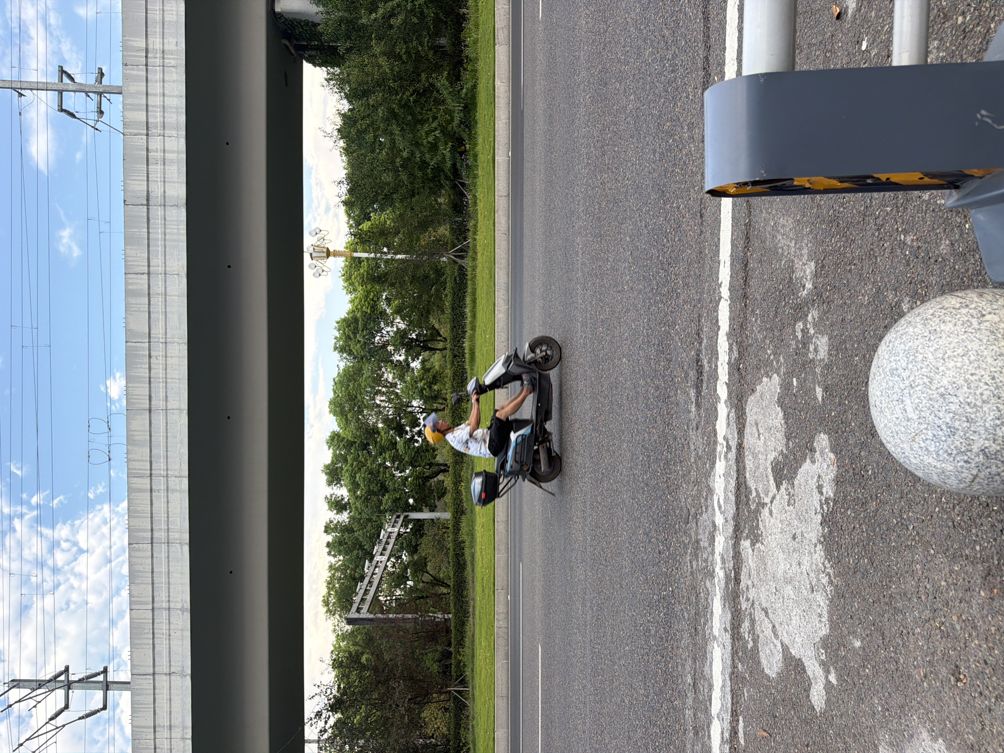 In Jiangjin, China, a person wearing a yellow helmet rides an electric scooter on a paved road. The scooter is black with a grey storage box on the back. The rider is wearing a light-colored short-sleeved shirt and dark shorts. The road is lined with a grassy median and trees. An elevated overpass with electrical wires runs across the top of the frame. In the foreground, part of a grey barrier and a granite sphere are visible. The sky is blue with scattered clouds.