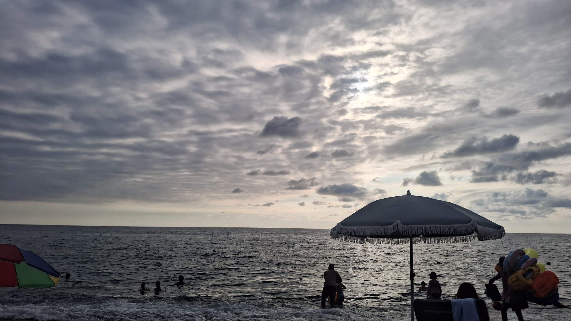 This image captures a coastal scene in Kobuleti, Georgia, likely on a cloudy day. The sea occupies the lower half of the frame, with gentle waves lapping at the shore. In the foreground, a large, dark umbrella with a fringed edge stands on the beach, casting a shadow. To its right, a person is seen selling brightly colored inflatable rings and toys. Several individuals are visible in the shallow water, some partially submerged, suggesting they are swimming or wading. In the distance, the horizon line separates the sea from the sky, which is filled with a textured layer of gray clouds, with patches of lighter sky and some sunlight peeking through. On the left side of the image, another colorful beach umbrella is partially visible. The overall atmosphere is one of a somewhat overcast but active seaside day.