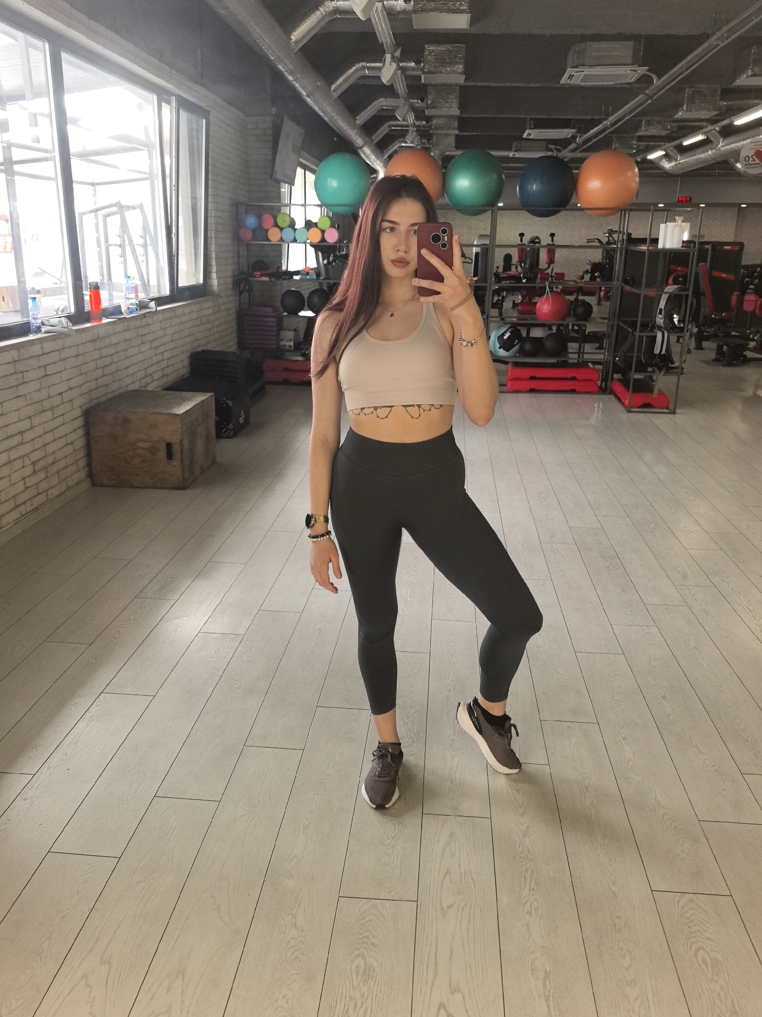 A young woman takes a selfie in a gym in Stara Zagora, Bulgaria. She stands on a light wood-grain floor with exercise equipment visible in the background. The gym has exercise balls, weights, and various machines. The woman wears a beige sports bra, black leggings, and athletic shoes. She holds a red smartphone up to take the picture. The gym also has a exposed ceiling with ducts. Large windows are along one wall.
