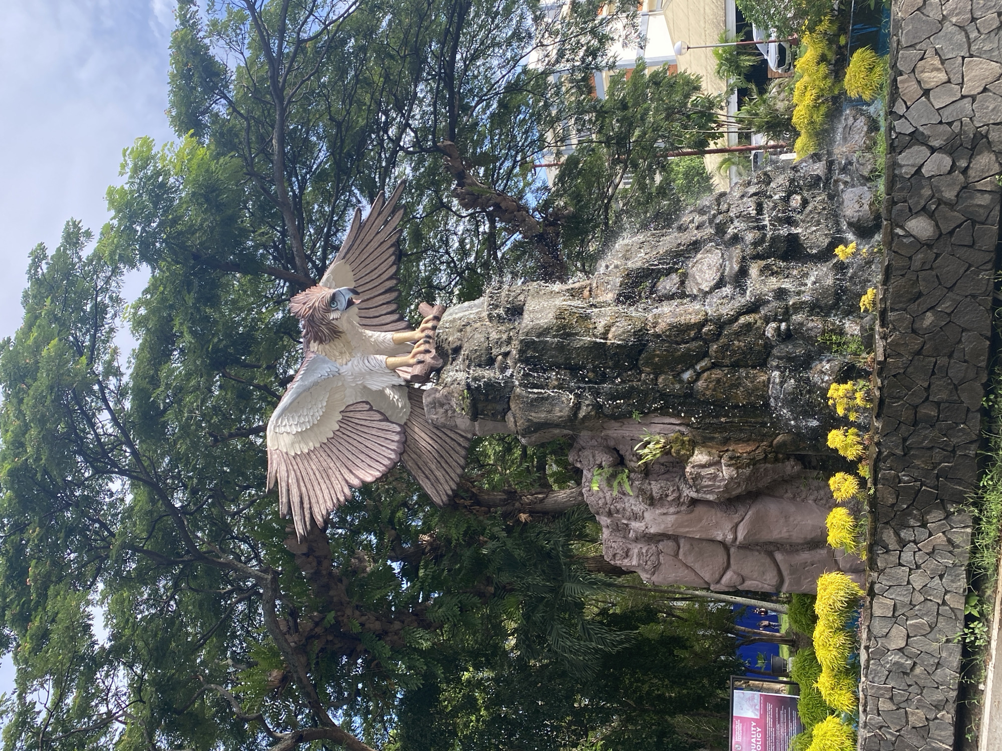 The image depicts a statue of a Philippine Eagle perched atop a stone waterfall feature in Davao City, Philippines. Water cascades down the rock structure. Green trees surround the statue and waterfall. A stone wall borders the right side of the image, adorned with yellow plants. A building is visible in the background. A sign is visible at the bottom of the image. The sky is partly cloudy.