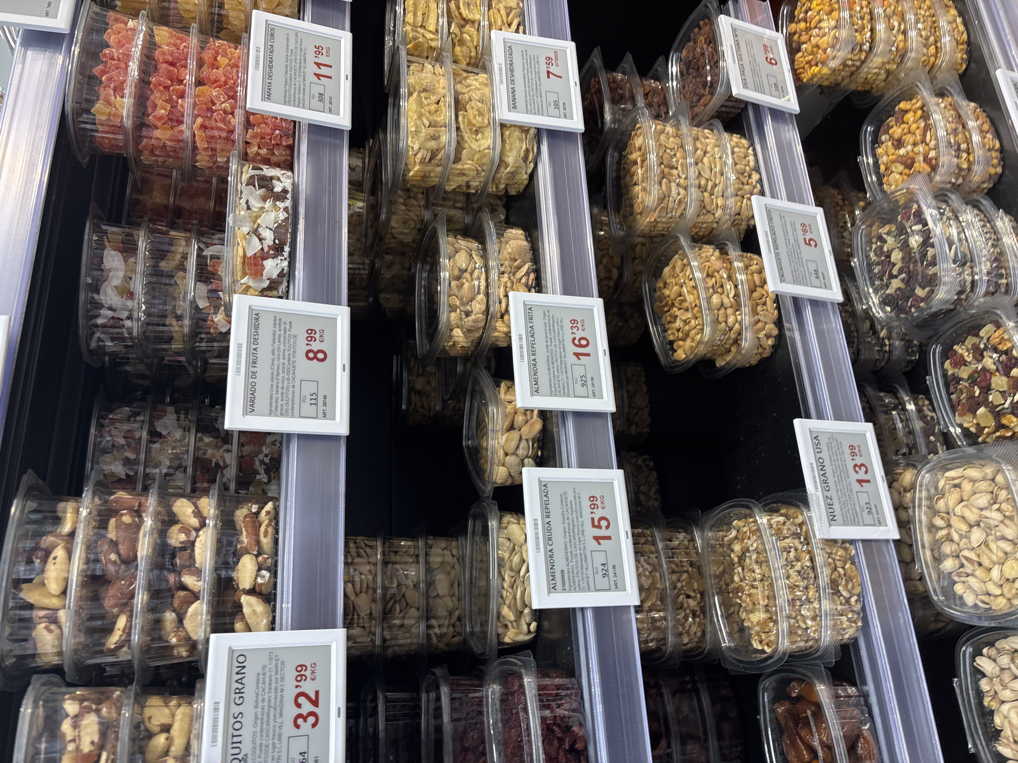 This is a full shot of a grocery store aisle in Urbanización Villa Martín, Spain. The aisle is filled with shelves displaying various packaged nuts and dried fruits in clear plastic containers. The products are stacked in multiple layers on the shelves, with price tags visible in front of some of them. The lighting appears to be artificial, typical of an indoor retail environment. There are no people or actions depicted in the image. The focus is on the arrangement and variety of food products.