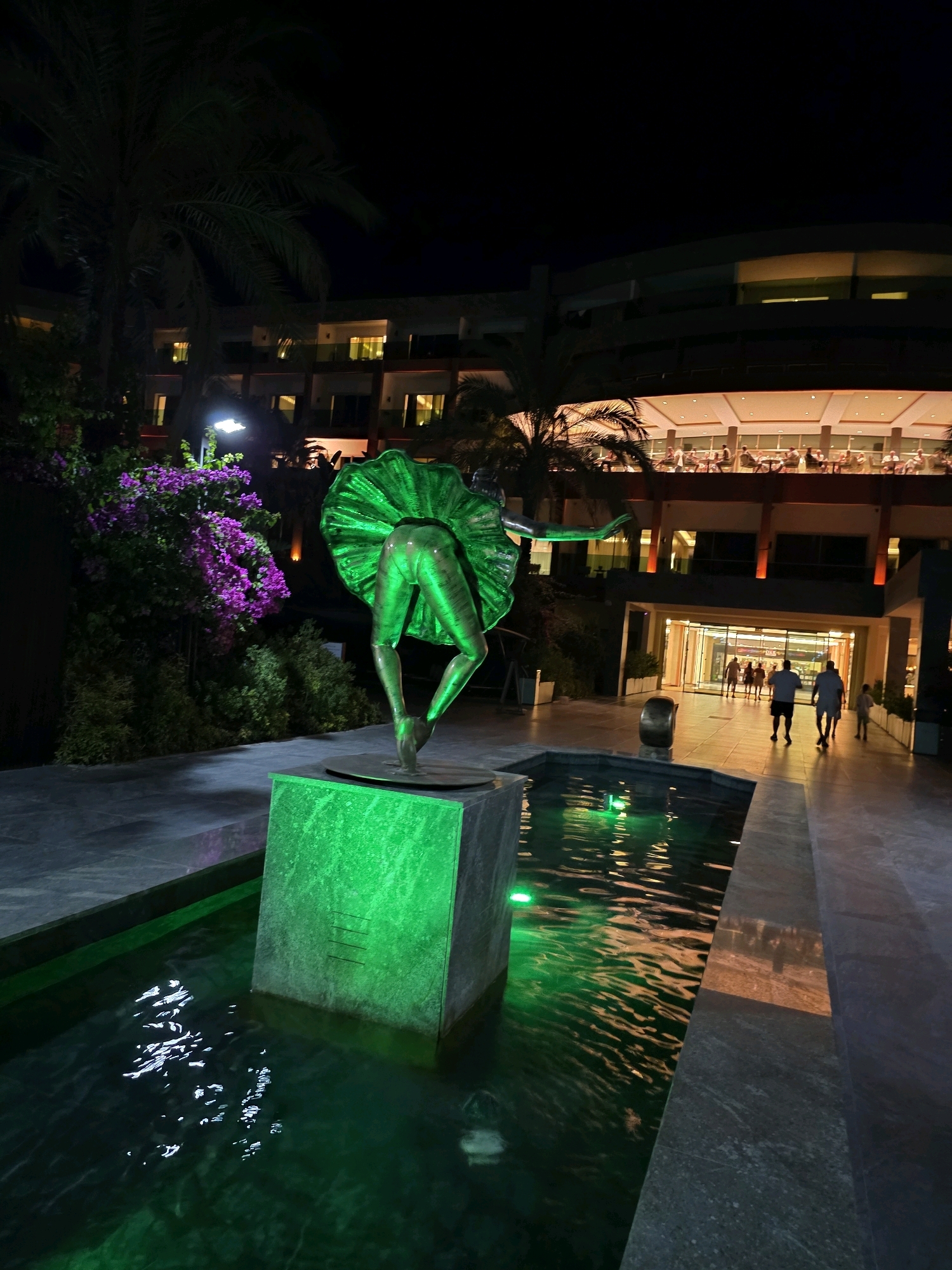 The image captures an outdoor scene at night in Bodrum, Turkey. In the foreground, a green-lit fountain features a bronze statue of a ballerina in mid-pose, her back to the viewer, with a voluminous skirt. The statue stands on a pedestal surrounded by water that reflects the green light. To the left of the fountain is a bush with vibrant purple flowers. Behind the fountain and statue, a modern building with multiple levels is visible, illuminated by interior lights and accent lighting on its facade. People are seen walking in the illuminated entryway of the building and seated on a balcony or terrace on an upper floor. Palm trees are present, suggesting a warm climate. The overall impression is a well-lit, contemporary resort or hotel setting at dusk or night.