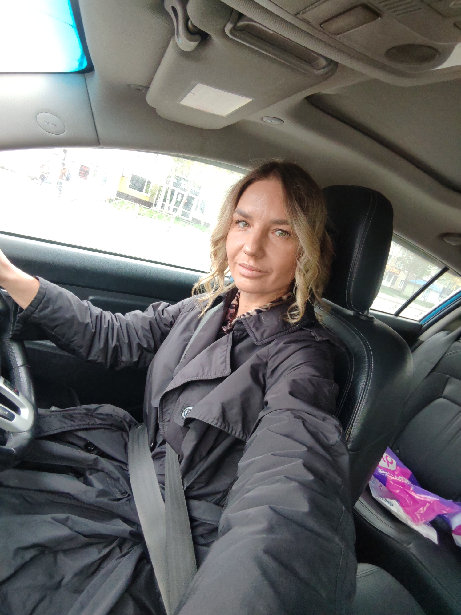 A woman with light brown wavy hair and green eyes is seen taking a selfie from the driver's seat of a car. She is looking directly at the camera with a calm, slightly smiling expression. She wears a dark, possibly black, long-sleeved jacket or trench coat over a patterned top, and a seatbelt is fastened across her chest and lap. Her hands are on the steering wheel, suggesting she is in the driver's position.
The car's interior features black leather seats and a grey headliner. A sun visor is visible on the passenger side, along with an "AIRBAG" label clearly printed on the A-pillar. The car appears to have a sunroof. On the rear passenger seat, a purple and white plastic bag or packaging can be seen.
Through the car windows, an outdoor scene is visible, depicting what appears to be an urban or suburban environment with buildings and trees, though specific details are somewhat blurry. The lighting suggests it is daytime, possibly overcast due to the diffused light and lack of harsh shadows. The overall setting implies the woman is either parked or briefly stopped in her car. The location context provided indicates this scene is in Petropavlovsk-Kamchatsky, Russia.