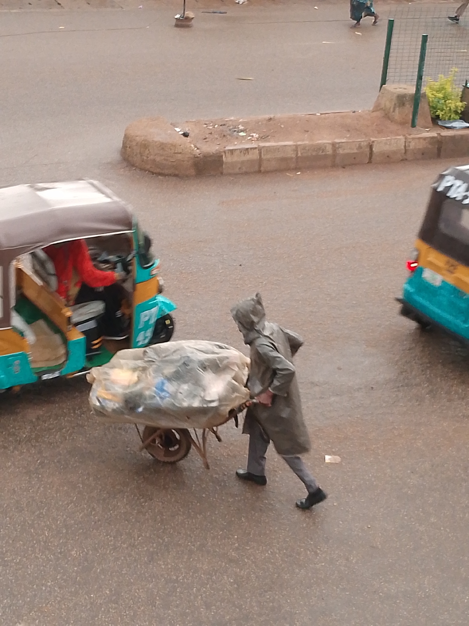In Bukuru, Nigeria, a person wearing a grey raincoat and hood pushes a wheelbarrow loaded with a large, clear plastic bag filled with an assortment of items, likely refuse, across a wet asphalt road. The wheelbarrow has a single wheel and metal handles. To the left, a teal and yellow auto-rickshaw with a brown canopy is stopped on the road, with a person in a red shirt visible inside. Further to the right, the rear of another auto-rickshaw, predominantly yellow and teal, is visible. The environment appears to be a street with concrete curbs and dirt patches. The ground is wet, suggesting recent rainfall.