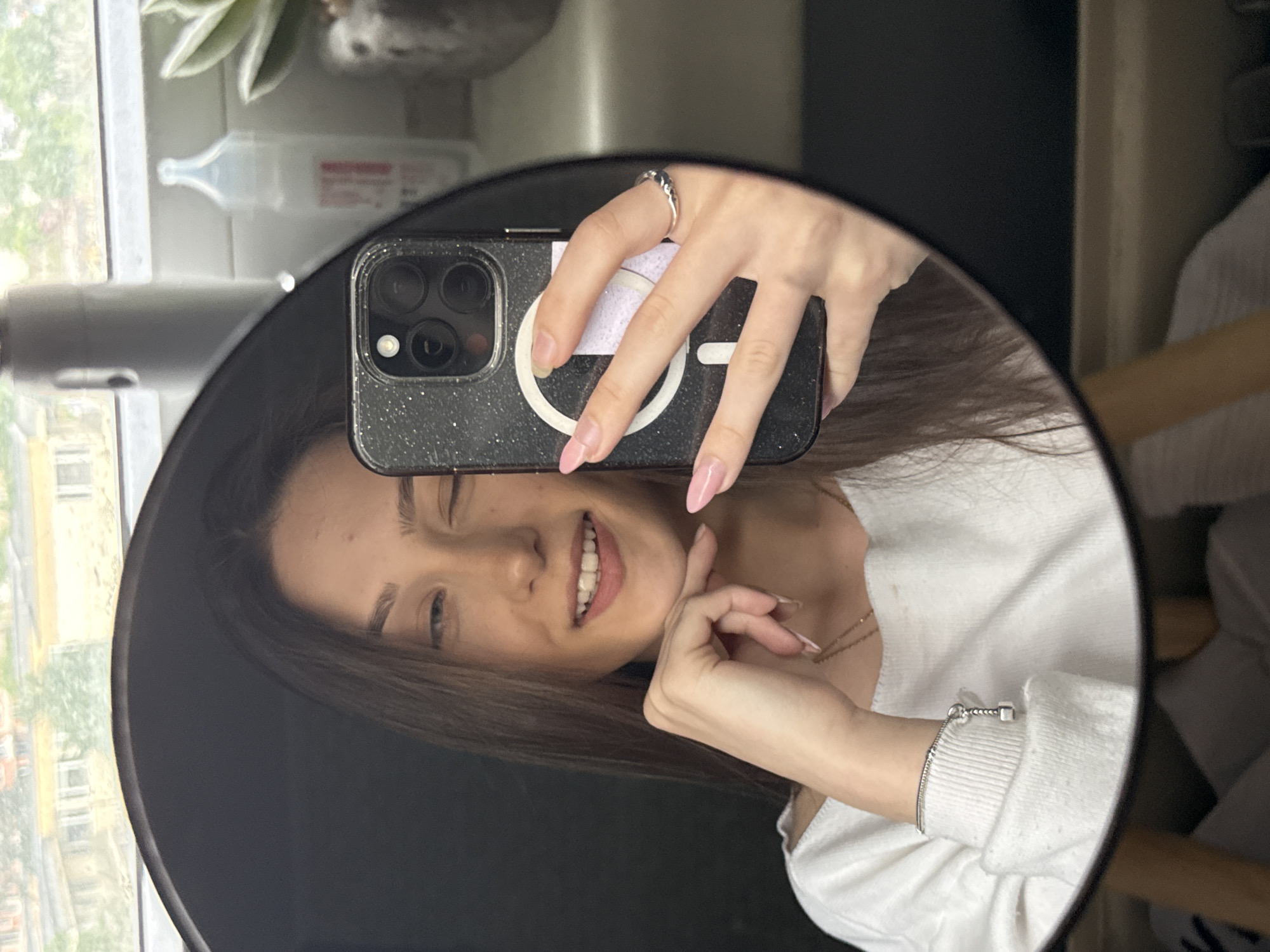 A young woman is taking a selfie in a round mirror. She is holding her phone up to the mirror with her left hand, partially obscuring her face. She has long brown hair and is wearing a white sweater. A ring is on her left hand. She is smiling. The phone has a black sparkly case with a white MagSafe charging circle. The background reflected in the mirror includes a window with a view of a city, a plant on a shelf, and a white bottle. A chair is visible on the right. The city location cannot be determined from the image.