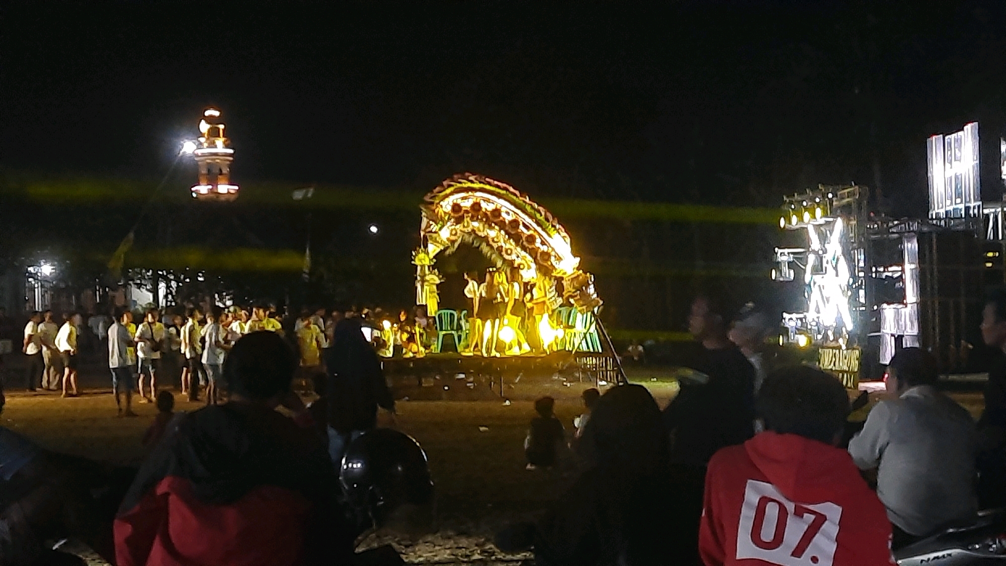 This image depicts an outdoor nighttime event in Karangdoro, Indonesia, possibly a festival or performance. A stage with performers, illuminated by bright yellow and white lights, is the central focus. The stage is decorated with a large, ornate archway resembling a dragon or traditional motif, also lit up. Several people are gathered around the stage, watching the performance. To the left, a tall, brightly lit structure resembling a minaret or tower stands against the dark sky. On the right side of the image, a large sound system setup with multiple speakers and lights is visible. The ground appears to be unpaved, with some debris scattered around. The overall atmosphere suggests a lively community gathering.