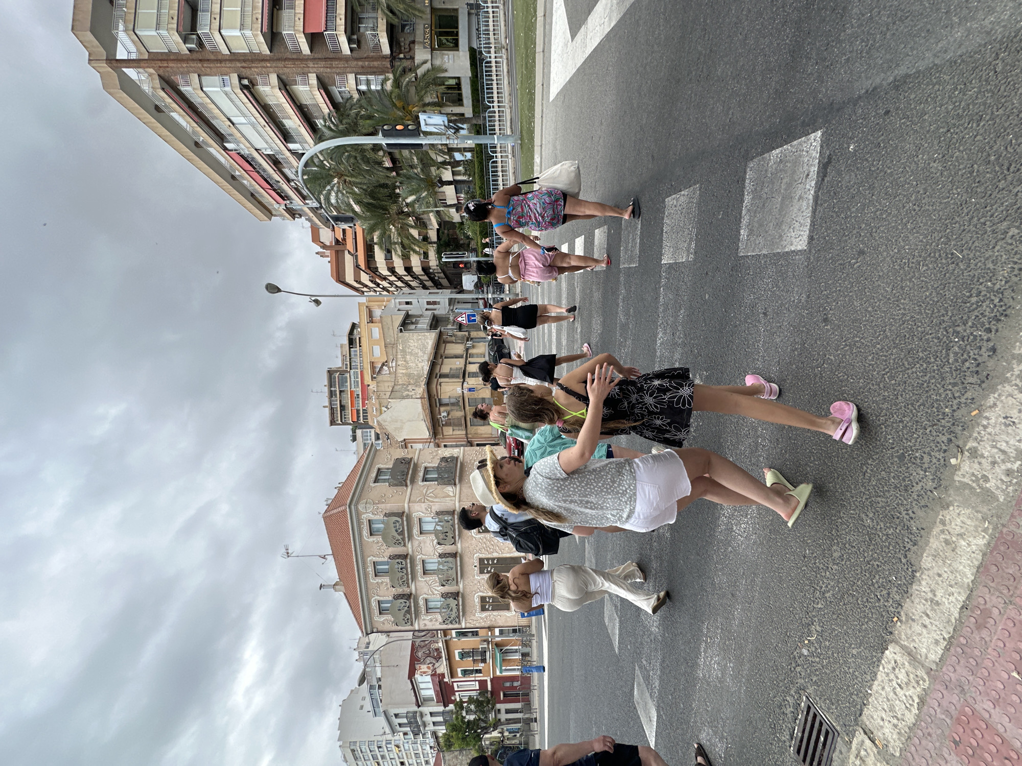In the city of Unknown, Unknown, a group of people are crossing a crosswalk on a cloudy day. The scene is urban, with buildings of various heights and architectural styles lining the street. In the foreground, two young girls are walking hand-in-hand across the asphalt, one wearing a black floral dress and pink Crocs, and the other in a white top, white shorts, and light green slides. They are surrounded by other pedestrians, some walking in the same direction, others in opposite.
Behind the girls, a woman in a straw hat and a patterned top, and a man with a backpack are also crossing. Further back, a woman in white pants and a strapless top is walking away from the camera. Other individuals are visible further down the crosswalk, including a woman in a pink dress and a patterned top. The road is marked with white stripes for the crosswalk, and a storm drain is visible on the left.
The background features several multi-story buildings, some with balconies and awnings. Palm trees are interspersed between the buildings and along the sidewalk. The sky is overcast with gray clouds, suggesting a possible change in weather. The overall impression is a bustling urban environment with people engaged in everyday activities.