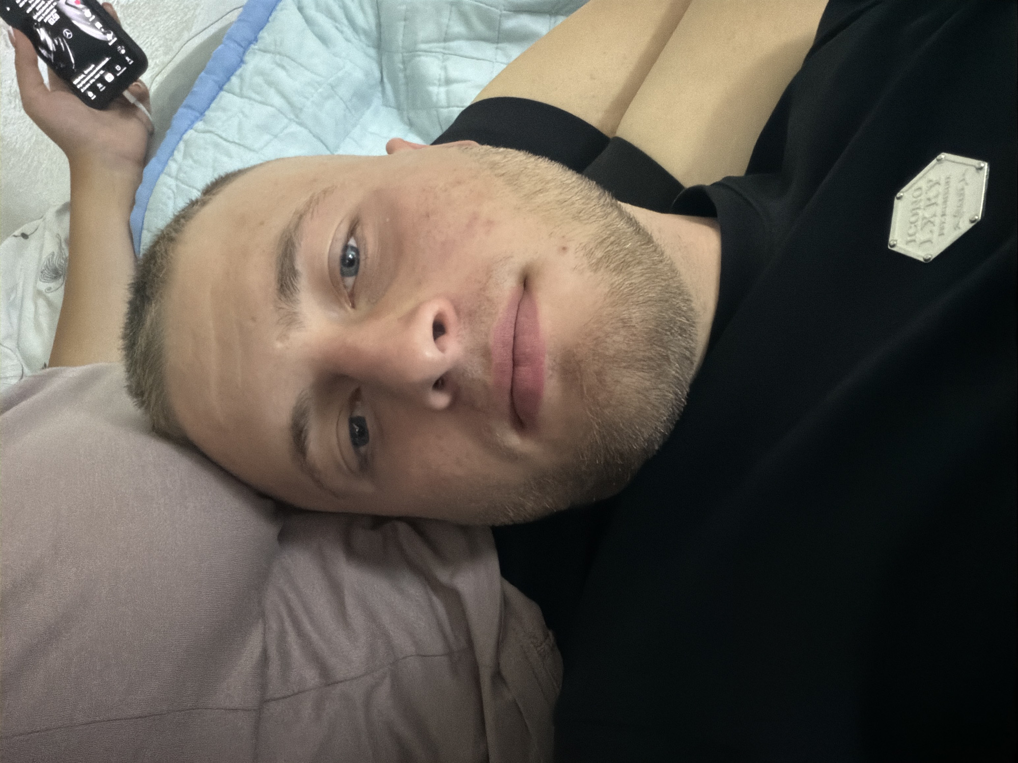 A close-up, high-angle, selfie-style photograph shows a man lying down, facing the camera. He has short, light brown hair and a beard. His eyes are open, and he appears to be looking at the camera. He is wearing a black t-shirt with a hexagonal silver emblem on the left side of his chest. To the left of the man's head, a light purple pillow is visible. Behind the man, a light blue quilted blanket is present. In the upper right corner of the image, a person's hand is holding a smartphone, displaying a screen with text and icons. The scene appears to be indoors, possibly a bedroom. There is no identifiable information within the image that places the scene in Strășeni, Moldova.