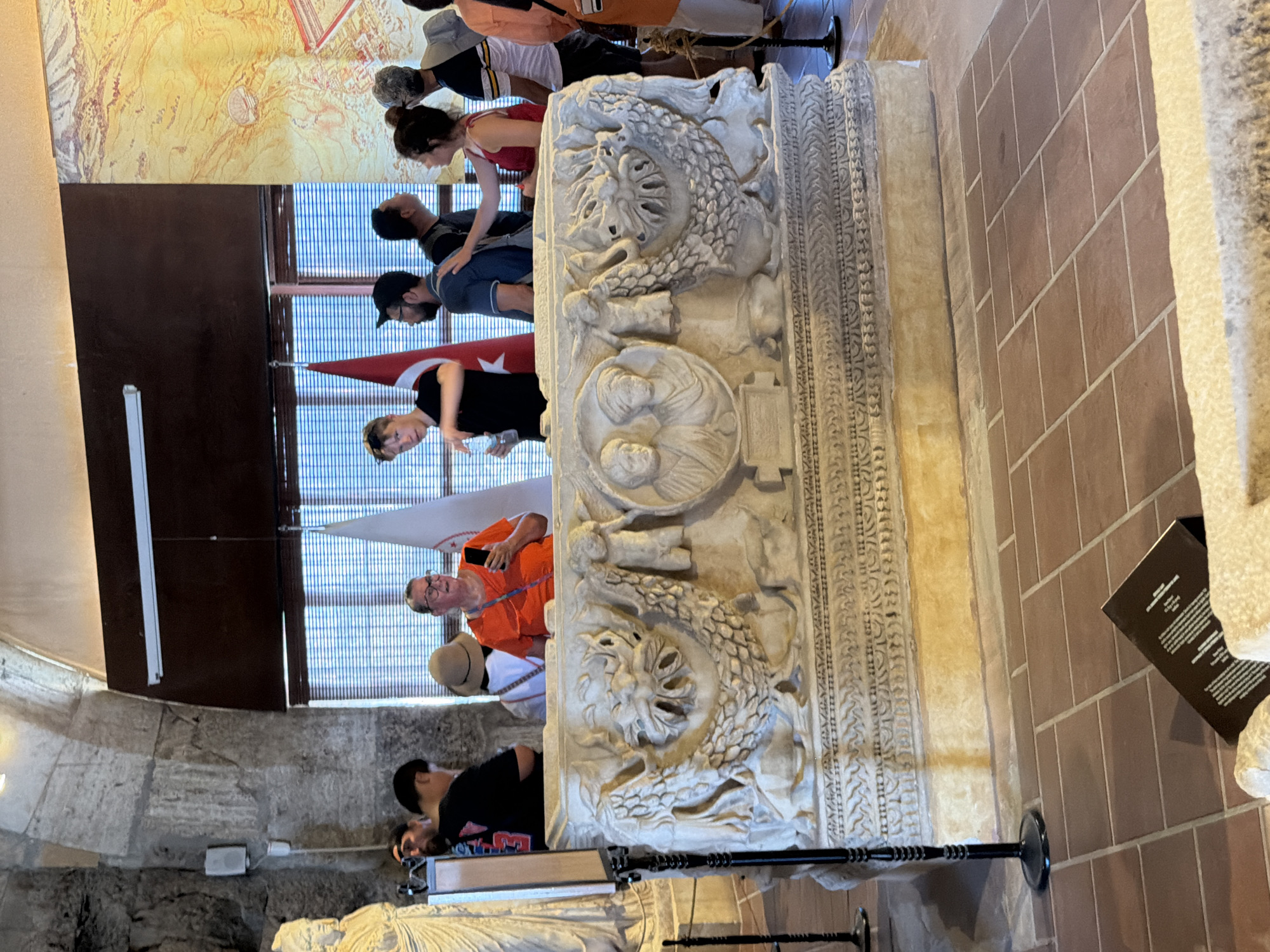 The image depicts the interior of a museum or historical site, possibly in Pamukkale, Turkey, given the context of such artifacts often found in the region. In the foreground, a large, ornate stone sarcophagus is prominently displayed. The sarcophagus features intricate carvings, including what appear to be dragon-like creatures on its sides and a circular relief with two faces on the front, flanked by angelic figures. The sarcophagus rests on a raised platform covered in yellow plaster. The floor is tiled with reddish-brown square tiles.
Several people are gathered around the sarcophagus, observing the artifact. In the mid-ground, a woman in an orange shirt is holding a mobile phone, looking at it. To her left, a young man in a black t-shirt holds a water bottle. Other individuals, men and women, are visible in the background, engaged in conversation or viewing the exhibits. Behind the sarcophagus, national flags, one red with a white crescent and star, are displayed. The architectural style of the building, with its arched ceilings and stone walls, suggests an ancient or historical structure. The background also includes a large, colorful mural or tapestry depicting a landscape.