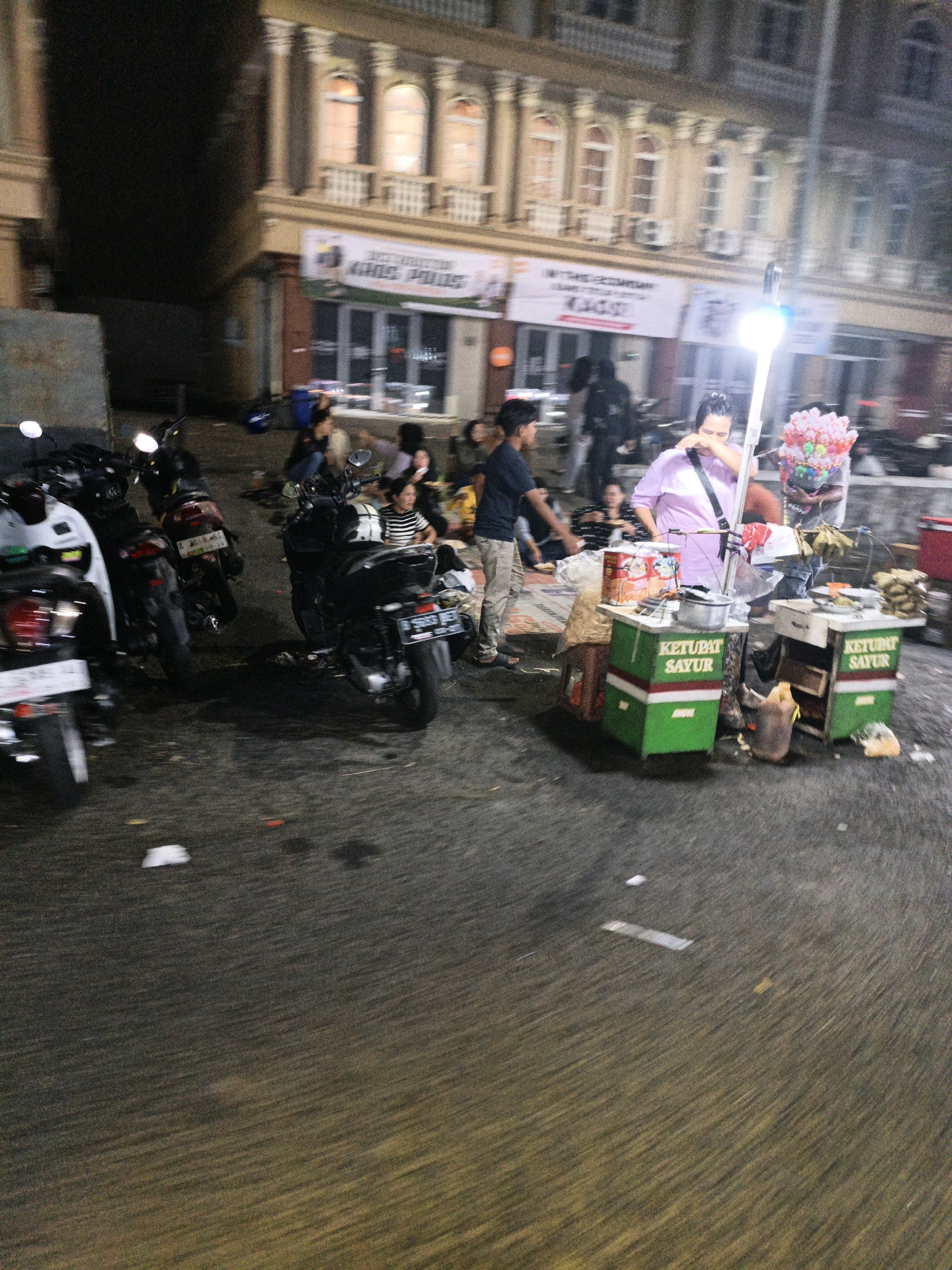 The image depicts a street scene at night in West Jakarta, Indonesia, likely a bustling area with a food vendor. A woman in a purple shirt stands behind a food cart selling "Ketupat Sayur," a traditional Indonesian dish. The cart is green and white, with branding visible. In front of the cart, there are bags of chips and other packaged food items. To the left of the cart, a man in a blue shirt and light-colored pants stands, seemingly interacting with someone. Several motorcycles are parked on the street, indicating a common mode of transportation in the area. In the background, a building with arched windows and balconies suggests a commercial or residential structure. The overall impression is that of a lively urban environment at night, centered around a food stall.