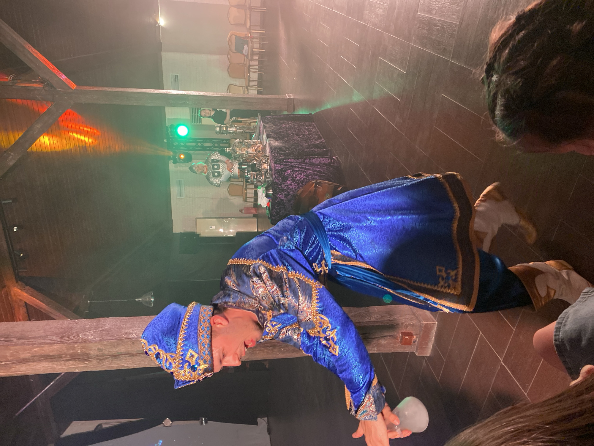 An indoor performance is captured in Abrau-Dyurso, Russia, illuminated by artificial lighting that suggests an evening event. The central figure is an entertainer, possibly a magician or theatrical performer, dressed in a vibrant royal blue costume richly embellished with intricate gold embroidery and sequin patterns. They wear a matching blue and gold hat or turban. The performer is posed dynamically, leaning over a large, weathered wooden beam, holding a glowing, translucent white spherical object in their hands, which appears to be a prop for their act.
In the background, a table covered with a dark, possibly purple velvet cloth holds several ornate objects resembling steampunk-inspired devices or trophies. Partially visible behind this table are other individuals, one wearing a black t-shirt, possibly staff or fellow performers. Stacked chairs are visible further in the distance. The setting includes rustic wooden beams overhead and to the left, indicating a venue with a distinct architectural style. Atmospheric lighting casts green and orange glows across the scene, with a clear green spotlight visible in the background. The floor is a dark-toned tiled or wooden surface arranged in a herringbone pattern. The heads of audience members are visible in the foreground and to the right, observing the performance. There is no discernible text visible in the image.