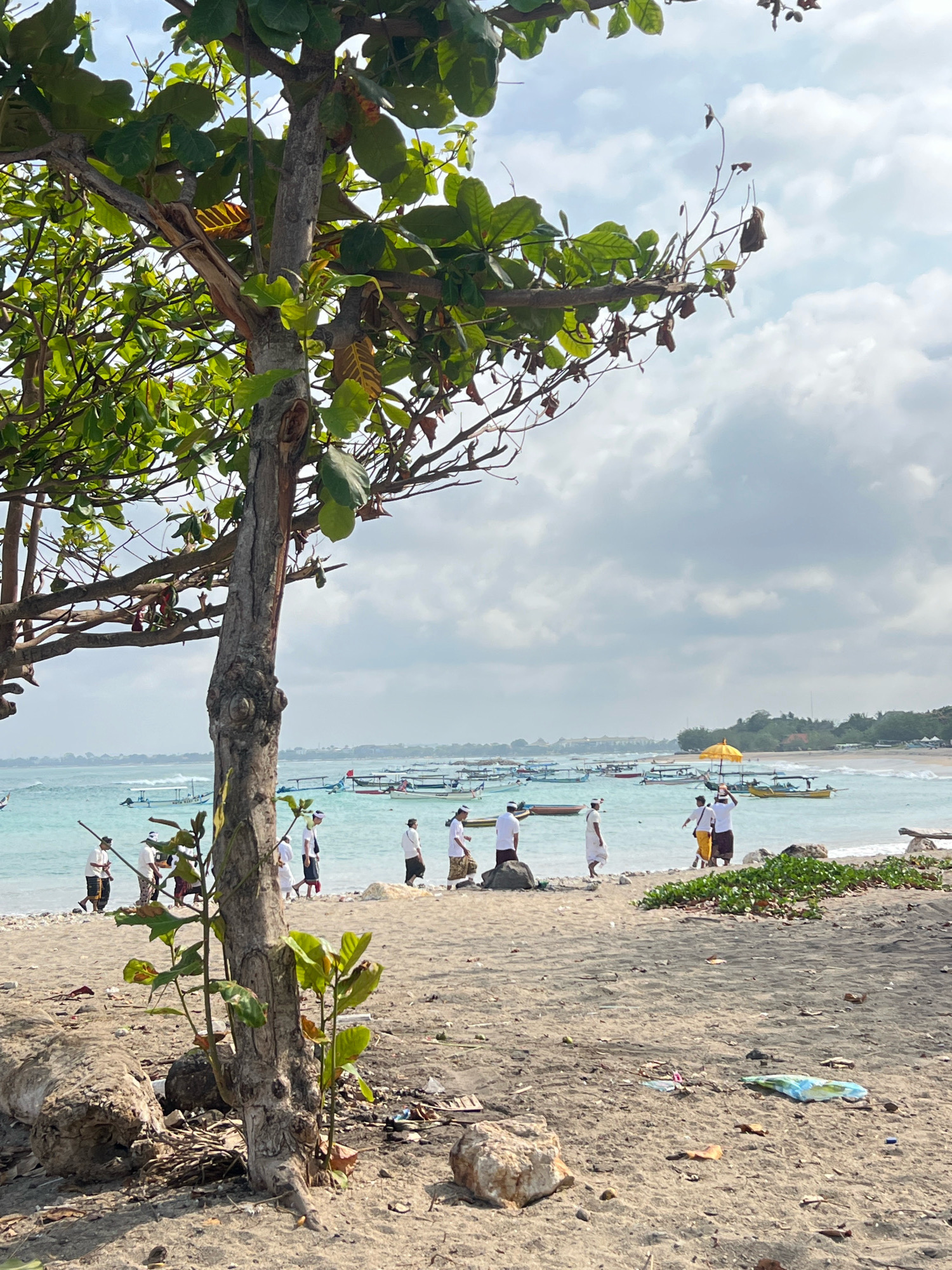 The image depicts a sandy beach scene in Kuta, Indonesia, with the ocean and several boats in the background. In the foreground, a tree with green leaves stands on the beach. Several people, some dressed in white attire, are walking along the shoreline. One person holds a yellow umbrella, and another carries an object on their head. The water is a clear turquoise, and the sky is filled with clouds. The beach is strewn with debris, including driftwood and plastic items. The overall atmosphere is of a coastal town on a cloudy day.