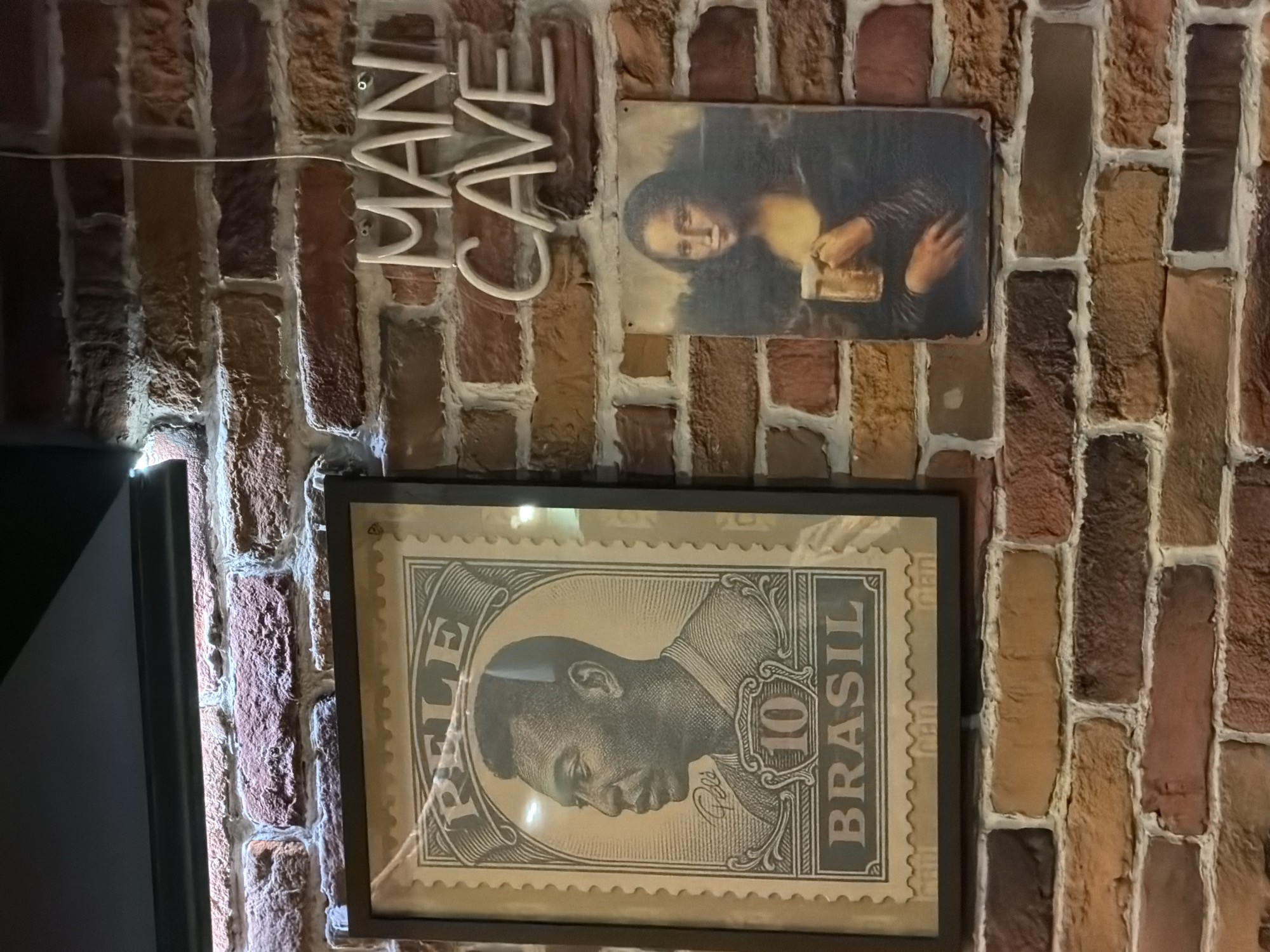 The image shows a brick wall with various decorations. There is a neon sign that says "MAN CAVE". Above a framed image of Pelé on a Brazilian stamp is a print of the Mona Lisa drinking a beer. The scene likely depicts an interior. There is insufficient information to determine the location in Sofia, Bulgaria.