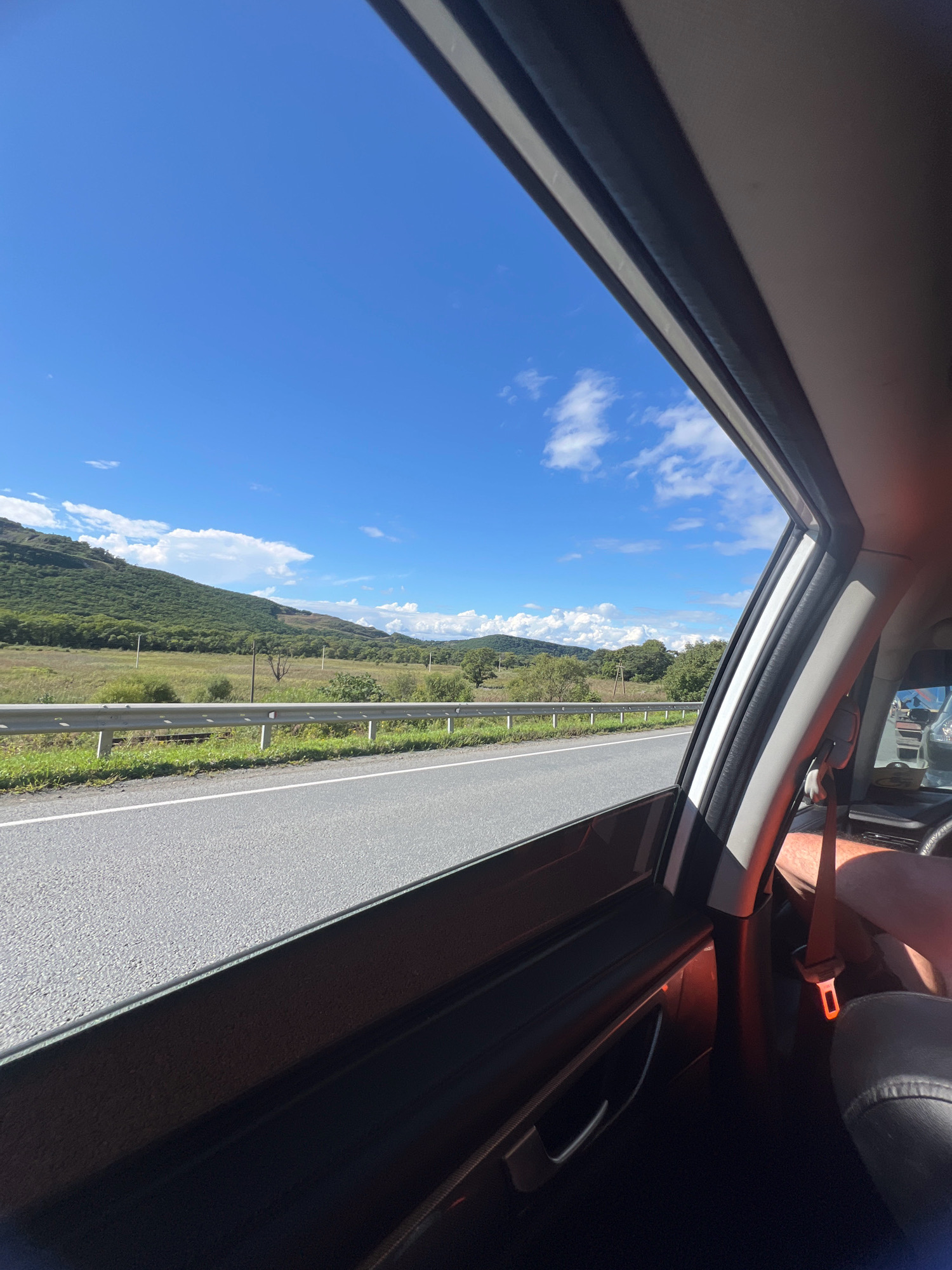 The image depicts a view from inside a vehicle looking out the window on a sunny day. The foreground is dominated by the interior of the car, showing a portion of the door panel, a seatbelt, and the edge of a car seat. Through the window, a paved road is visible, bordered by a metal guardrail and a grassy verge. Beyond the road, a field of dry grass stretches towards a gentle, tree-covered slope. The sky is a vibrant blue with scattered white clouds. The scene appears to be a rural or semi-rural setting, with no discernable urban features suggesting it is not located in the city of Bolshoy Kamen, Russia.