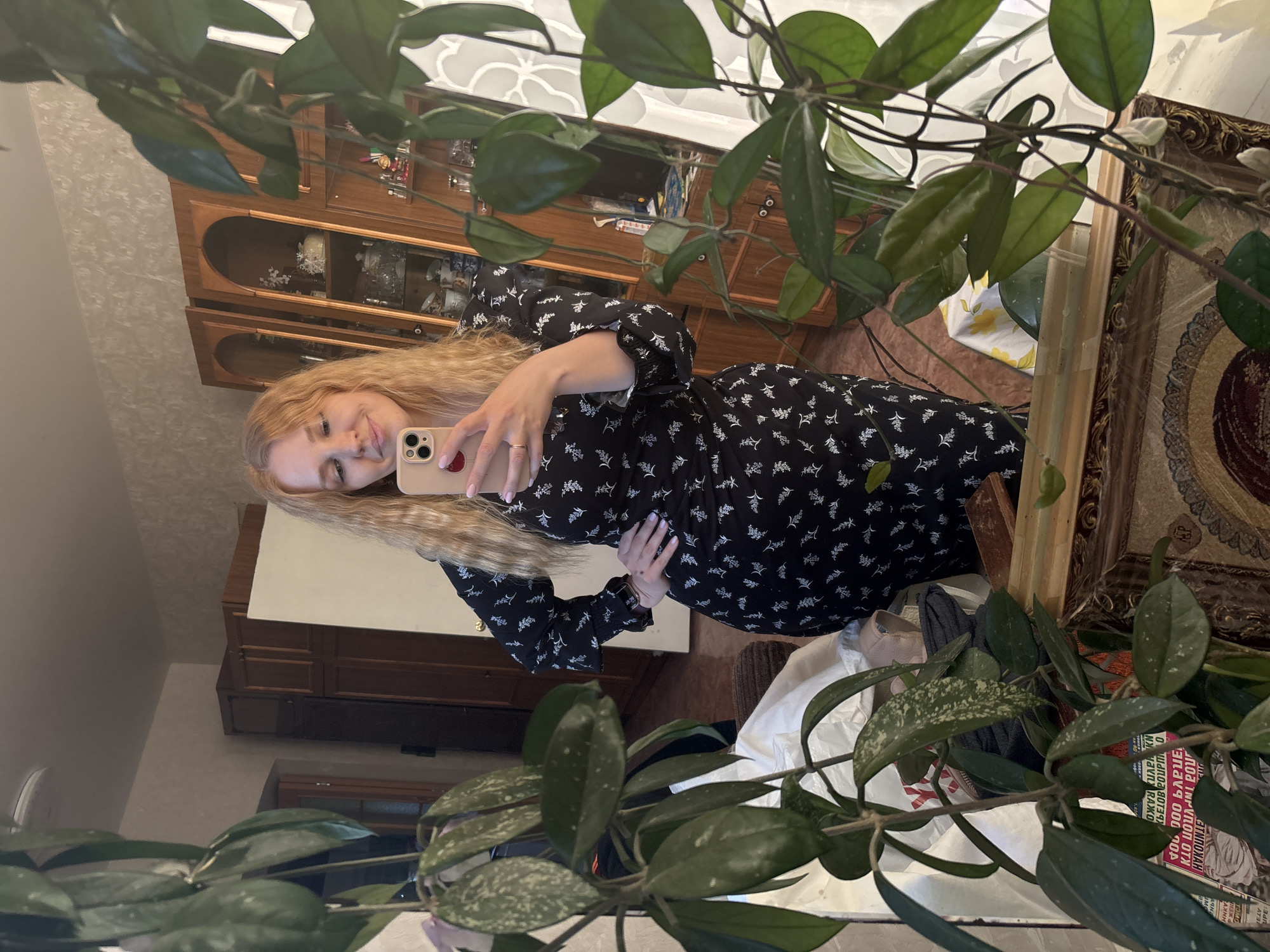 In Rozhdestveno, Russia, a female subject is depicted in a mirror selfie, partially obscured by a large, variegated leafy houseplant. The subject, with long, wavy blonde hair, is wearing a black long-sleeved dress adorned with a white floral pattern. She holds a light-colored smartphone, equipped with a multi-lens camera array, in her right hand, which is positioned in front of her chest. Her left hand rests on her hip. She has light-colored nail polish and a ring on her left ring finger. The mirror's lower section features an ornate, possibly gilded, frame. Reflected in the mirror is a domestic interior featuring light-colored, patterned wallpaper. The background includes two tall, wooden display cabinets with glass doors and arched tops, containing various ceramic and glass decorative items. To the left, a darker wooden wardrobe is visible. A light-colored ceiling with a circular fixture is reflected above. At the bottom of the reflection, a wooden floor and a surface covered by a yellow and white floral tablecloth are visible. Items near the bottom of the mirror include a white bag and papers with Cyrillic text.