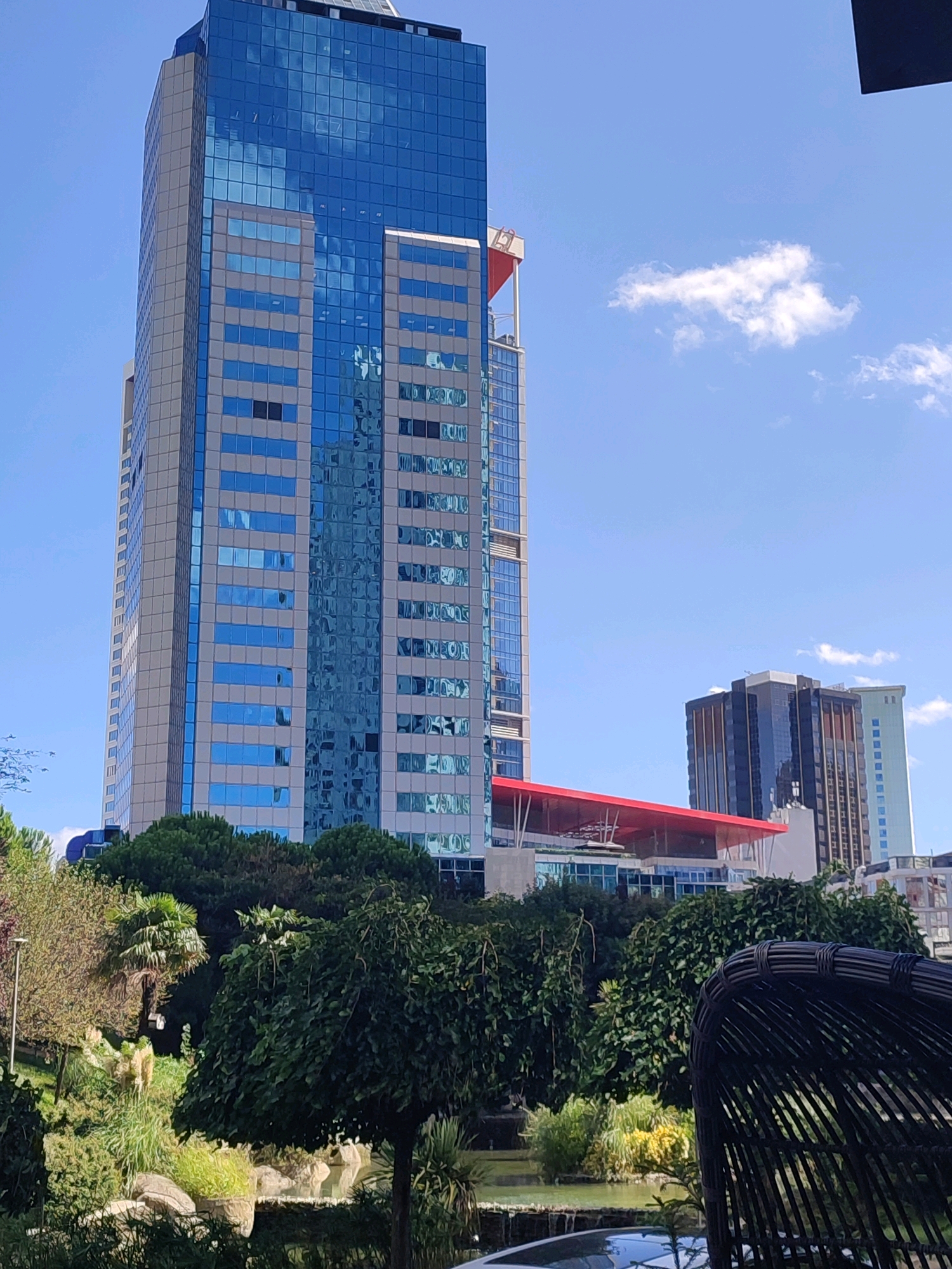 A tall, modern skyscraper with a blue glass facade dominates the scene, set against a clear blue sky with scattered white clouds. To the right of the main skyscraper, two other buildings are visible, one with a dark glass exterior and vertical gold accents, and another lighter colored building. In the foreground, a lush green park area with various trees, bushes, and a small pond is depicted. A portion of a dark wicker chair is visible in the lower right corner. This image appears to be taken in the city of Sarıyer, Turkey, likely in a commercial or business district. There are no people visible in the frame.
