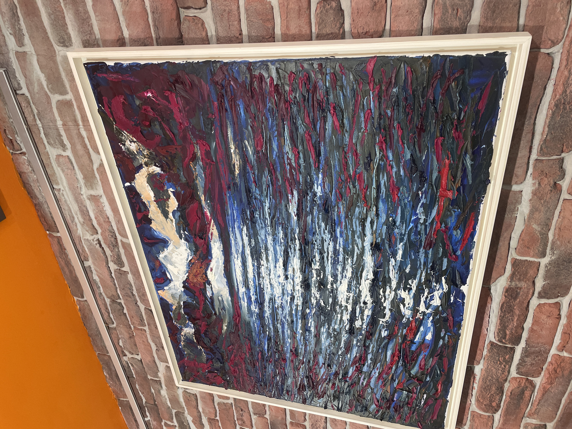 A thickly textured, abstract oil painting of a seascape is displayed on a brick wall. The painting features bold strokes of dark blues, blacks, and vibrant reds, with streaks of white and lighter blues suggesting choppy water reflecting light. The sky portion of the painting is a turbulent mix of deep reds, purples, and whites, with hints of orange suggesting a dramatic sunset or sunrise. The painting is framed in a light-colored, simple frame. Above the painting, an orange ceiling or soffit is visible. The brick wall has a rough, rustic appearance with varying shades of red and brown bricks and light-colored mortar. The scene is illuminated by artificial light, creating shadows and highlighting the impasto technique of the artwork. The location is stated as being in the city of Burgas, Bulgaria, although specific details within the image do not confirm this location.