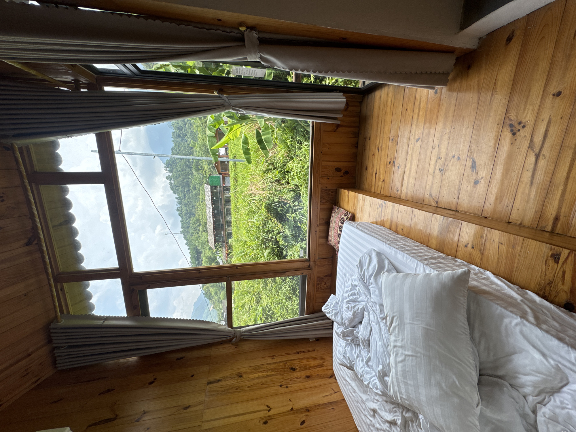 The image displays an interior room, likely a guesthouse or lodge, with a large window offering a panoramic view of a lush, mountainous landscape. The room itself features prominent wooden elements, including a warm-toned wooden floor with visible grain and knots, and matching wooden paneling on the walls.
On the right side of the frame, a bed with white, striped sheets and pillows is partially visible, appearing comfortably unmade. Adjacent to the bed and beneath the window is a wooden bench or platform, adorned with a small, colorful patterned throw pillow. Light-colored curtains are tied back, allowing maximum natural light to enter and providing an unobstructed view of the outdoors.
Outside the large, multi-paned wooden window, a vibrant natural scene unfolds. Dense, verdant vegetation covers rolling hills, with distant mountains visible under a partly cloudy sky. Distinct banana plants can be seen among the foliage closer to the window. A faint glimpse of a structure, possibly another building, is visible in the middle distance.
The bright daylight streaming into the room indicates it is daytime. The overall atmosphere conveyed is one of rustic tranquility and a deep connection to nature, fitting for the given context of Du Già Commune, Vietnam. There is no visible text in the image.