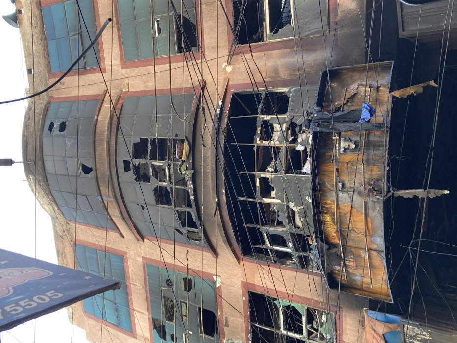 This image depicts the facade of a severely fire-damaged, multi-story building in Quetta City Tehsil, Pakistan, likely in a commercial or mixed-use urban area. The building, constructed of a light brown/tan material, exhibits extensive charring and soot stains across its lower visible floors. Numerous windows on multiple levels are shattered or completely missing glass panes, exposing darkened interiors with visible debris.
A prominent, curved metal balcony on the lower portion of the visible structure is heavily rusted, discolored by heat, and exhibits material degradation, including peeling surfaces. Two dark fabric items, possibly articles of clothing (a dark garment and a piece of blue fabric), hang over the railing of this damaged balcony.
Electrical wires are visible crisscrossing irregularly in front of the building. A partially visible blue sign with white text, including the numbers "5505," is attached to the upper-left side of the building. No individuals are present in the scene. The sky above is bright and overcast.