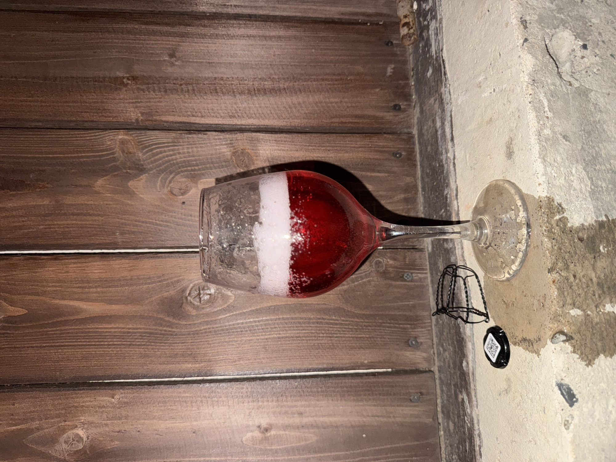 A wine glass filled with a red sparkling beverage sits on a concrete surface in front of a wooden wall. A black bottle cap with a QR code and a wire cage from a champagne bottle lie on the concrete near the glass. The scene is dimly lit, with the wooden wall providing a rustic backdrop. The location is identified as Chaikovsky, Russia. There are no people visible in the image, and no actions or interactions are depicted.